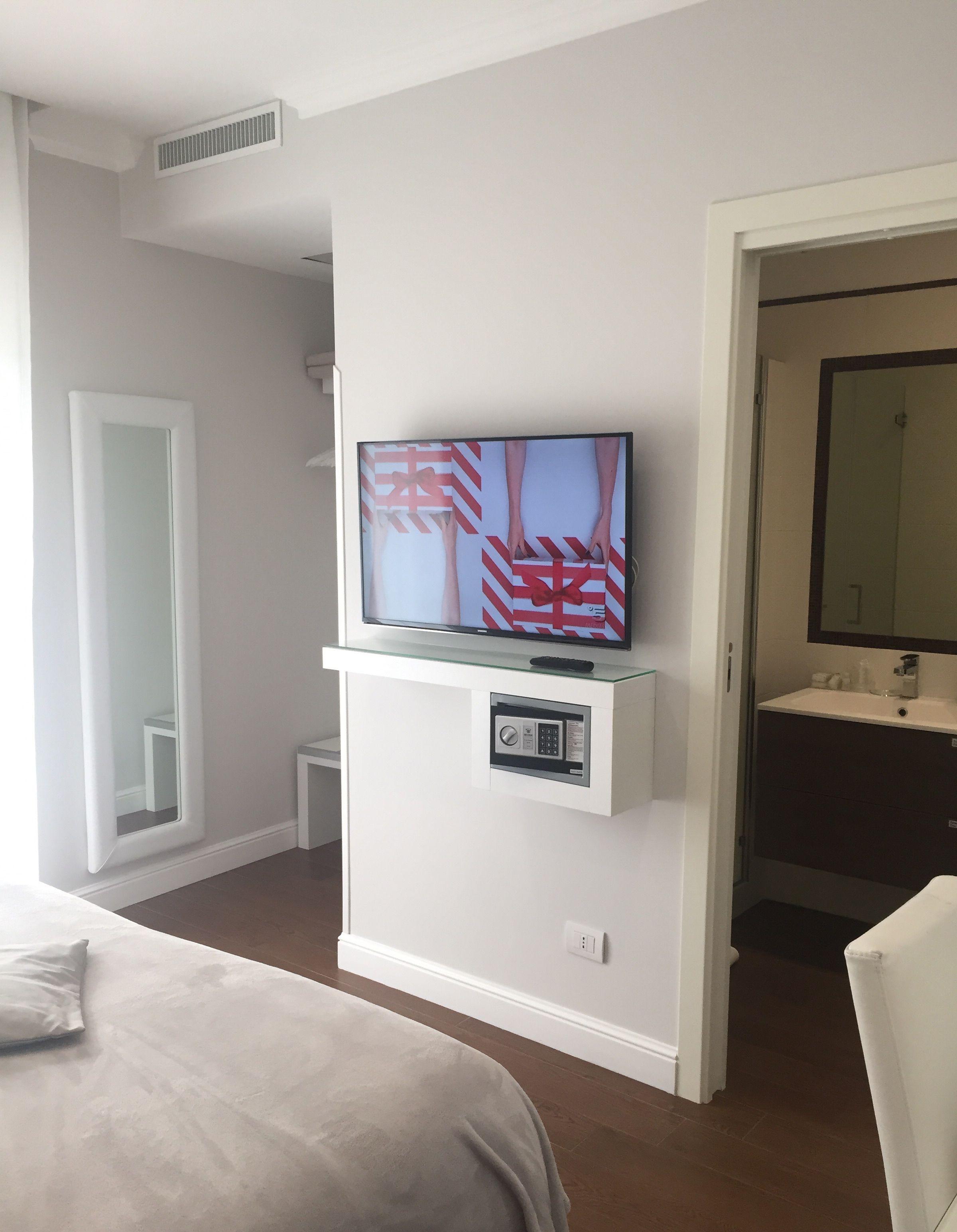 Hotellihuone, jossa on sänky, lasihyllylle asennettu televisio, pieni yöpöytä, peili sekä kylpyhuone, jossa on pesuallas ja peili.