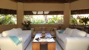 A room with two white sofas, a wooden coffee table, and a view of tropical plants through large windows.