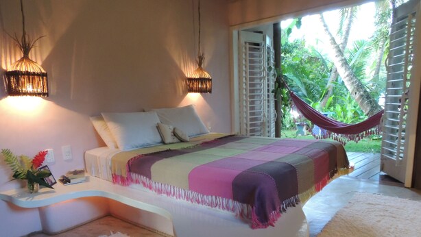 A bedroom with a bed, bedside lamps, a nightstand, a window with a view of palm trees, and a hammock.
