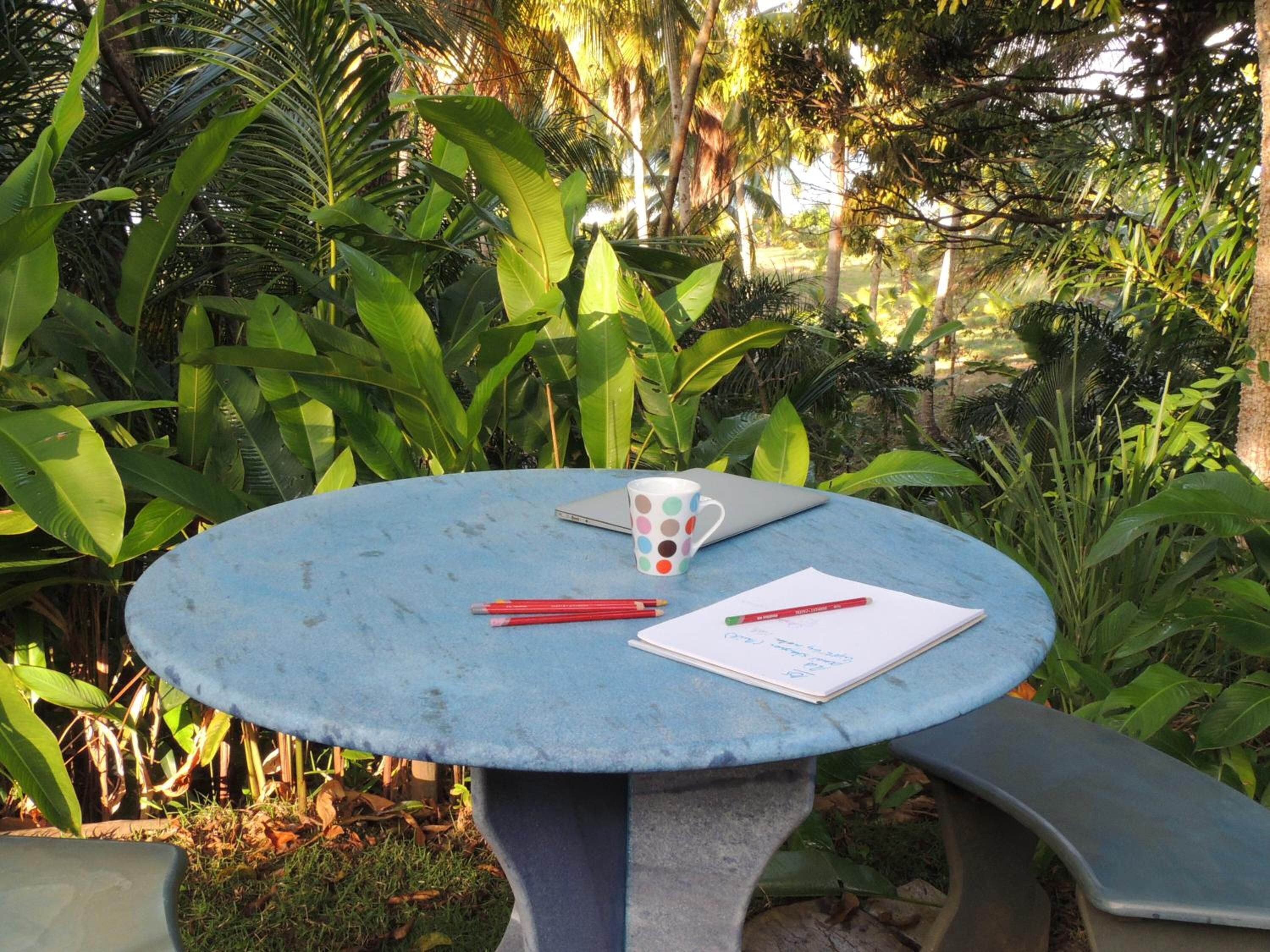 Una mesa redonda con una computadora portátil, una taza de lunares y un cuaderno con un bolígrafo rojo.