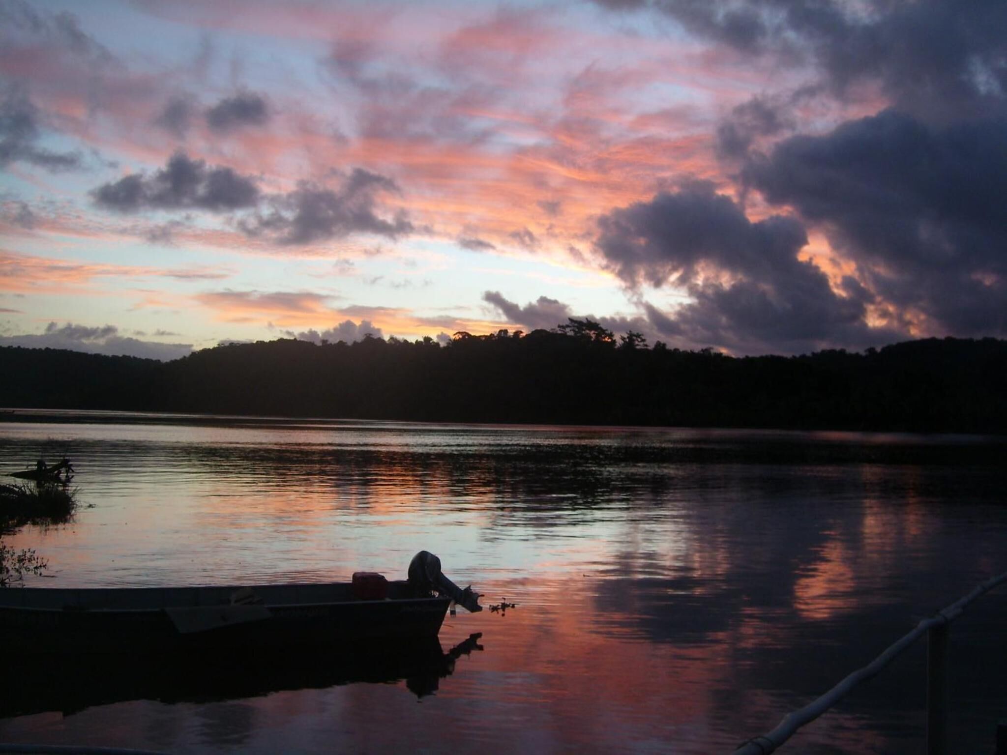 A boat docked at a calm lake during sunset with a forested shoreline in the background.