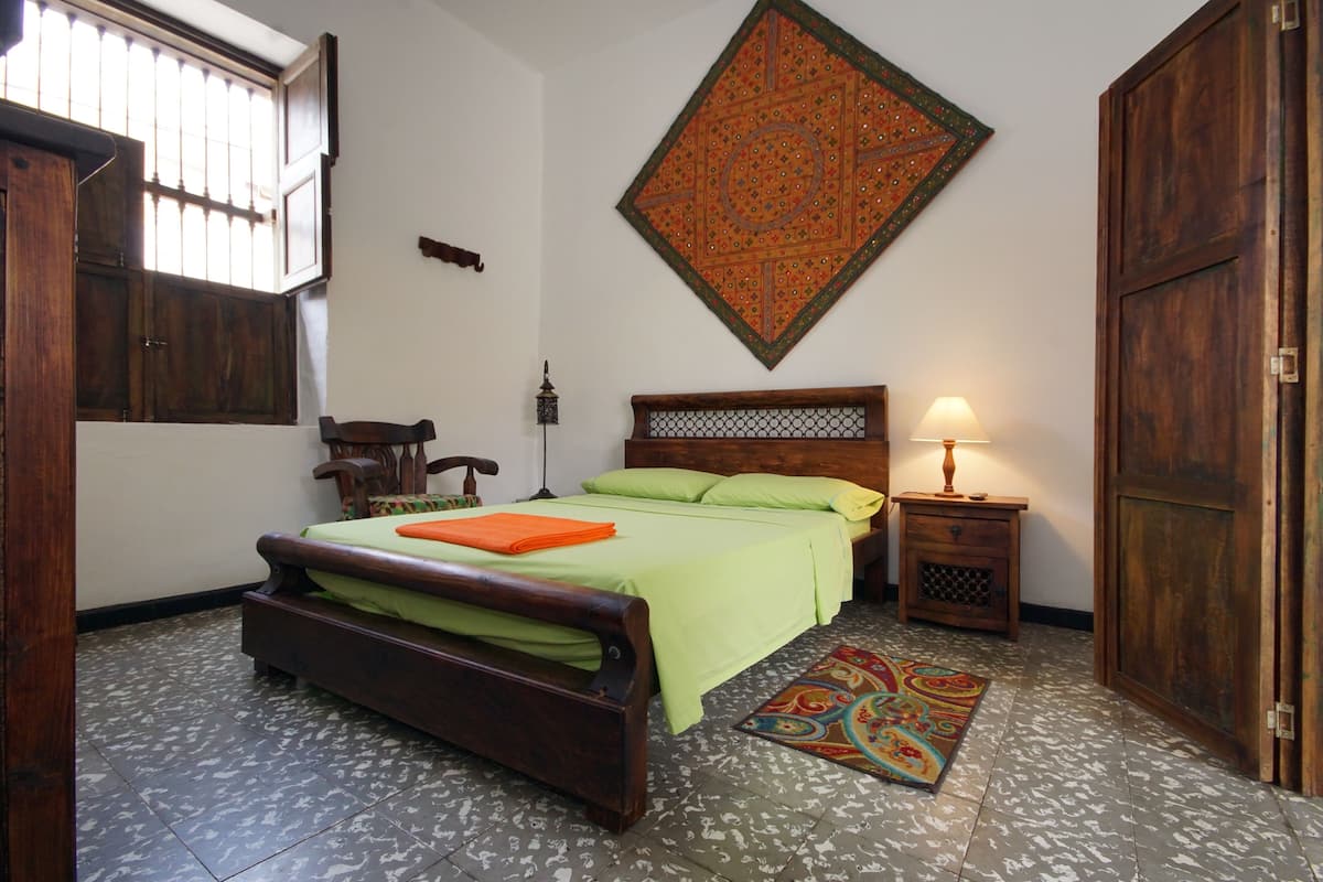 A room with a wooden bed, a nightstand with a lamp, a colorful rug, and a large decorative wall hanging.
