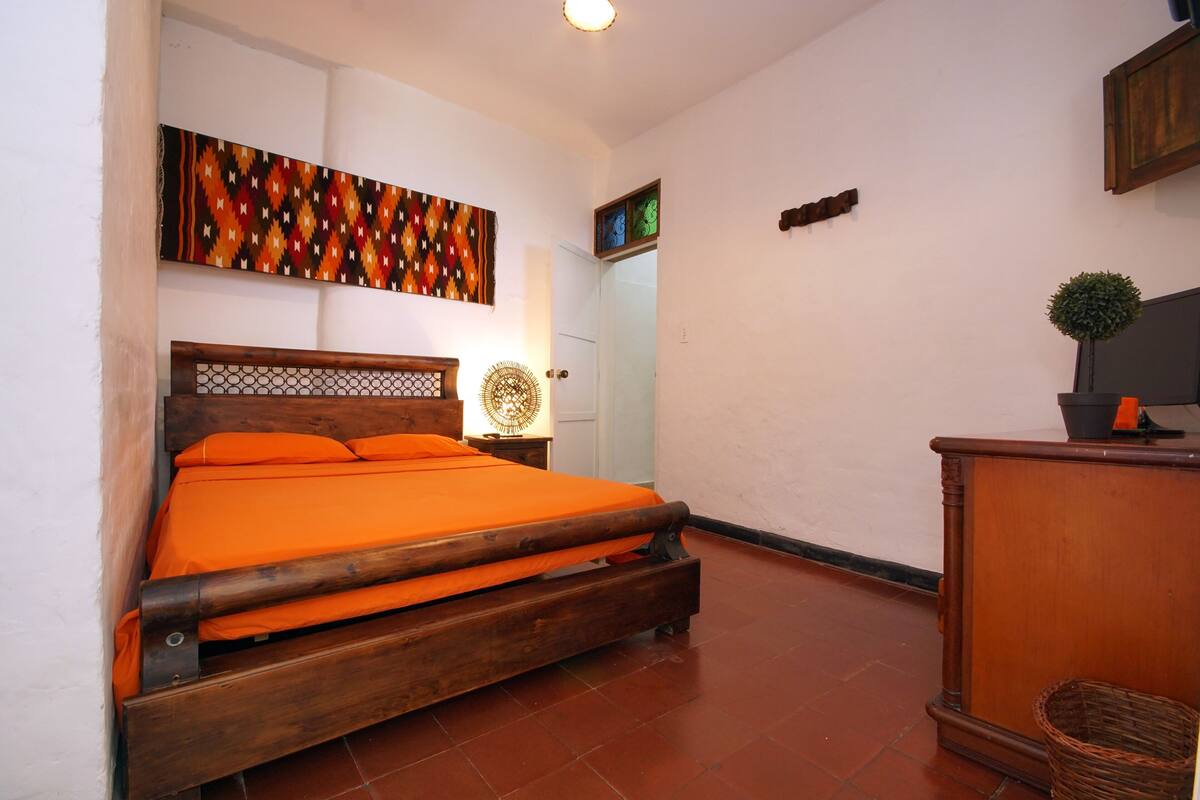A bedroom with a wooden bed, an orange bedspread, a wall-mounted artwork, a small table with a lamp, and a wooden dresser.