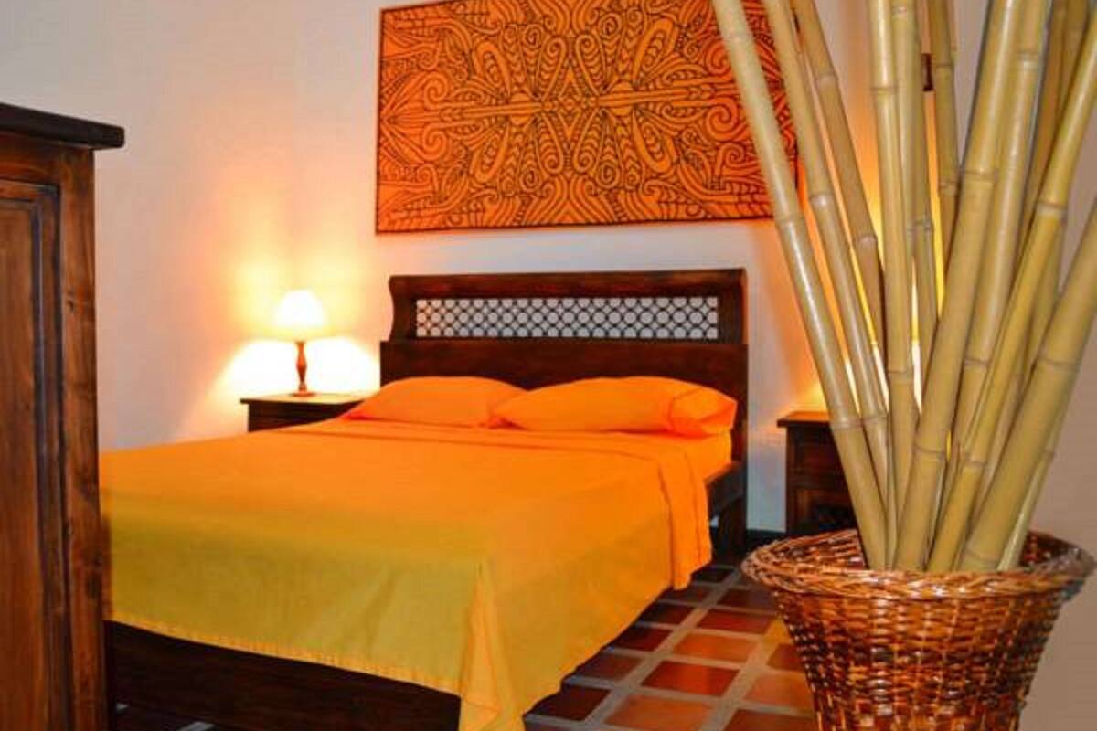 A room with a bed, a nightstand, a lamp, a wall hanging, and a bamboo plant.