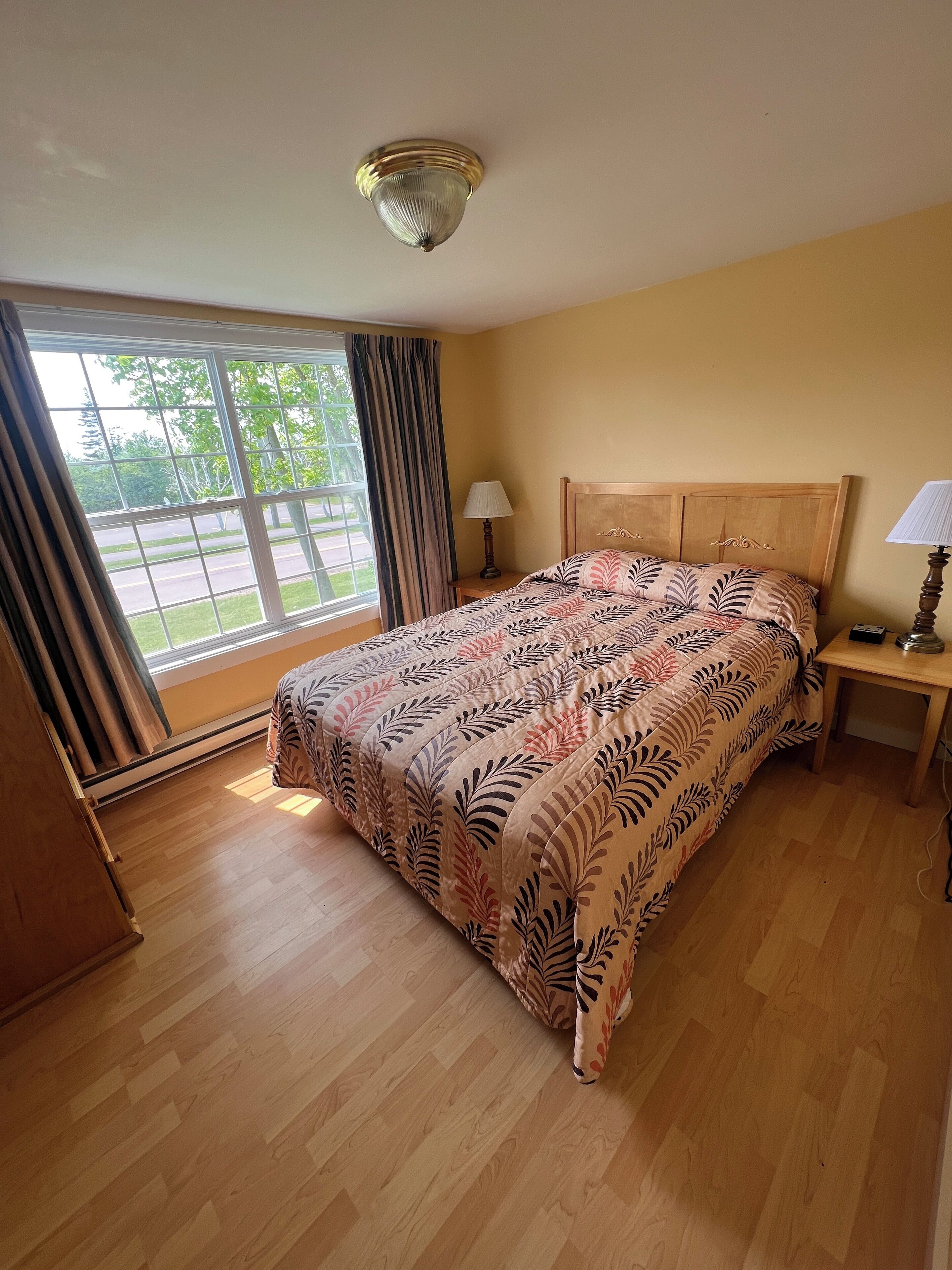 A hotel room with a large bed, wooden headboard, a nightstand with a lamp, a window with curtains, and a view of the outdoors.