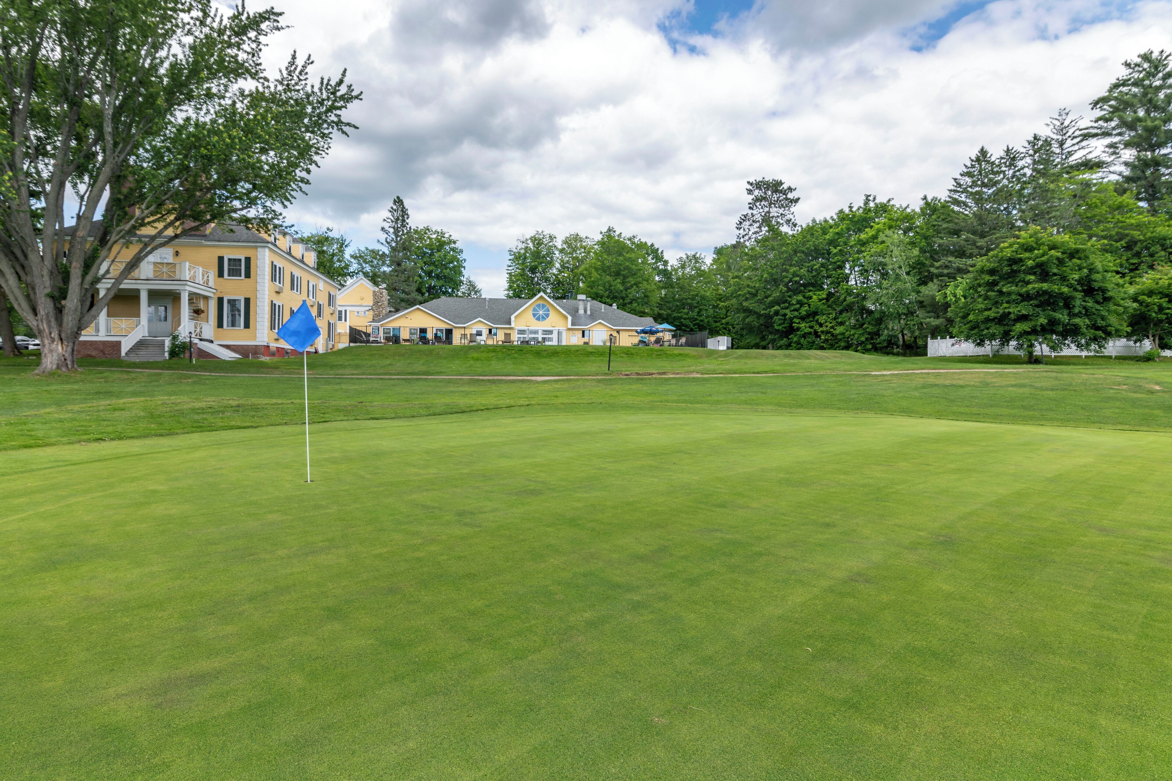 A golf course with a flagstick, a building with a yellow facade, and surrounding trees.