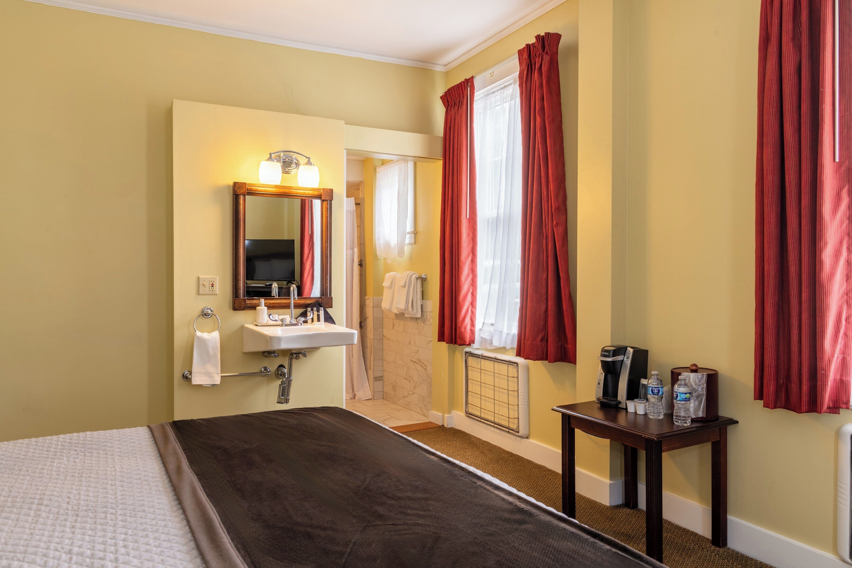A hotel room with a bed, a bathroom with a sink, mirror, and towel rack, a small table with toiletries, and a window with red curtains.
