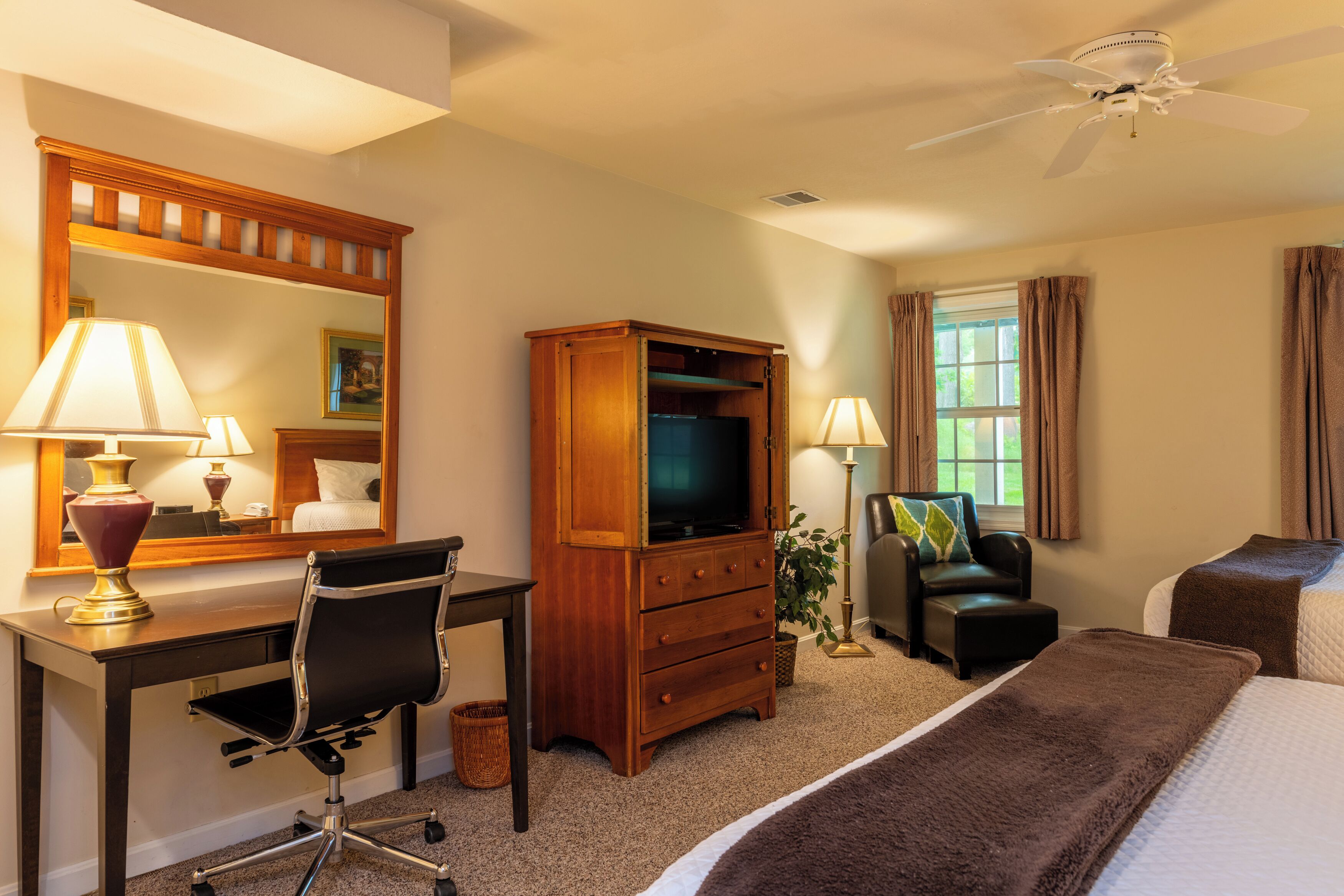 Een hotelkamer met een bureau, stoel, televisie, dressoir, bed en een plafondventilator.