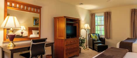A hotel room with a desk, chair, television, dresser, bed, and a ceiling fan.
