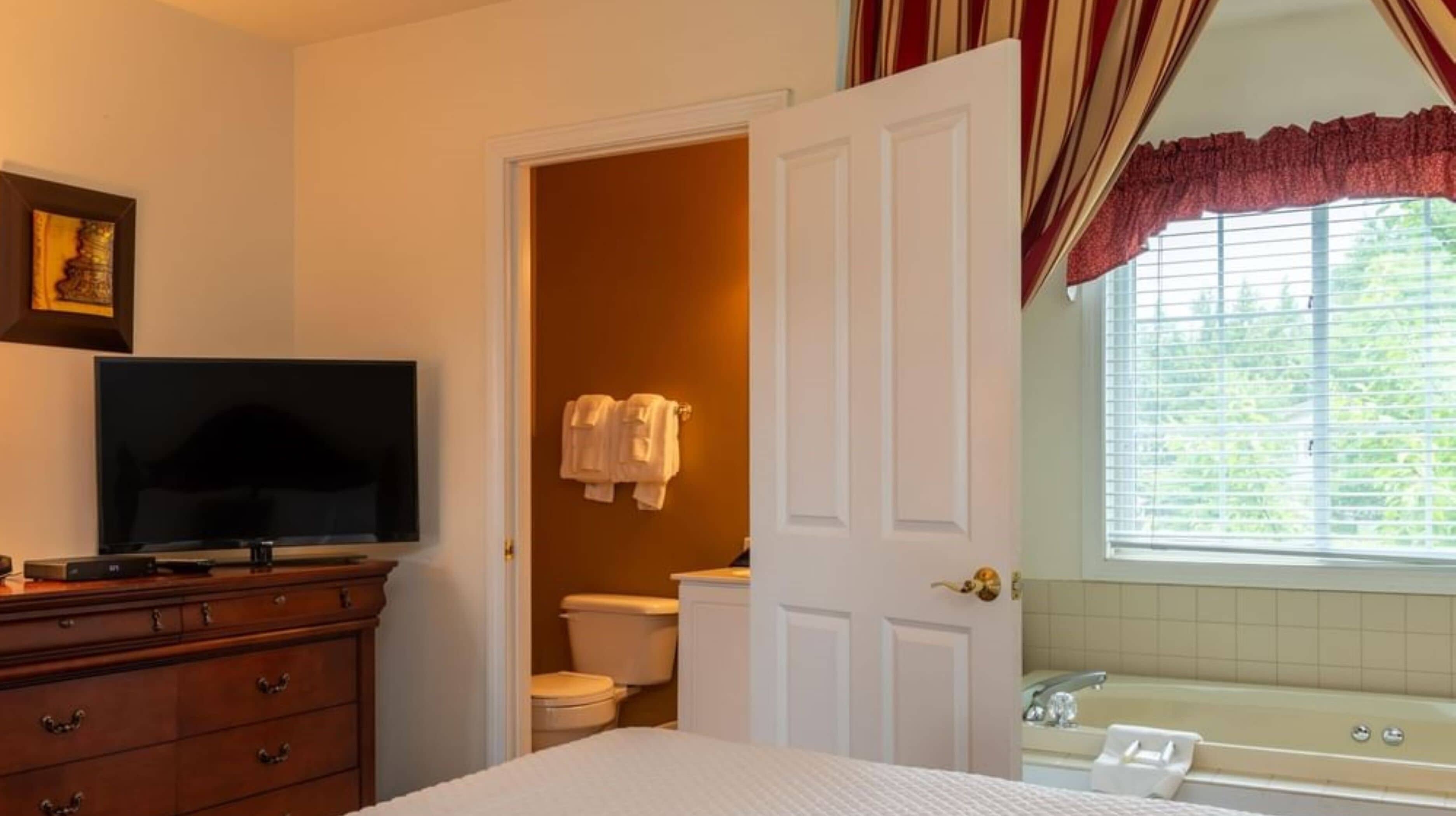 A bedroom with a bed, a dresser, a television, a bathroom with a toilet, a bathtub, and a window with curtains.