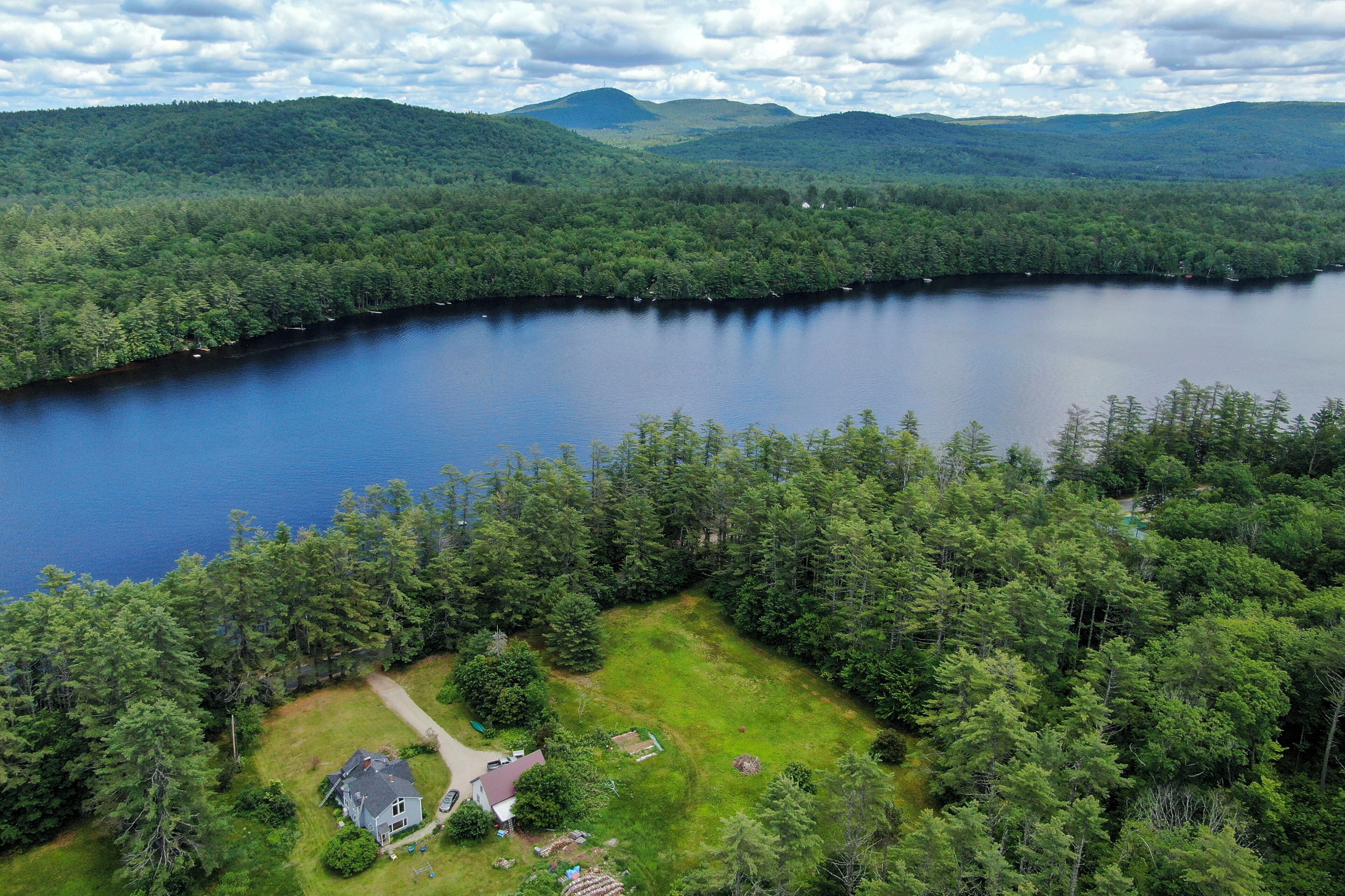 A lakeside property with a house surrounded by dense forest, a clear view of the lake, and rolling hills in the background.