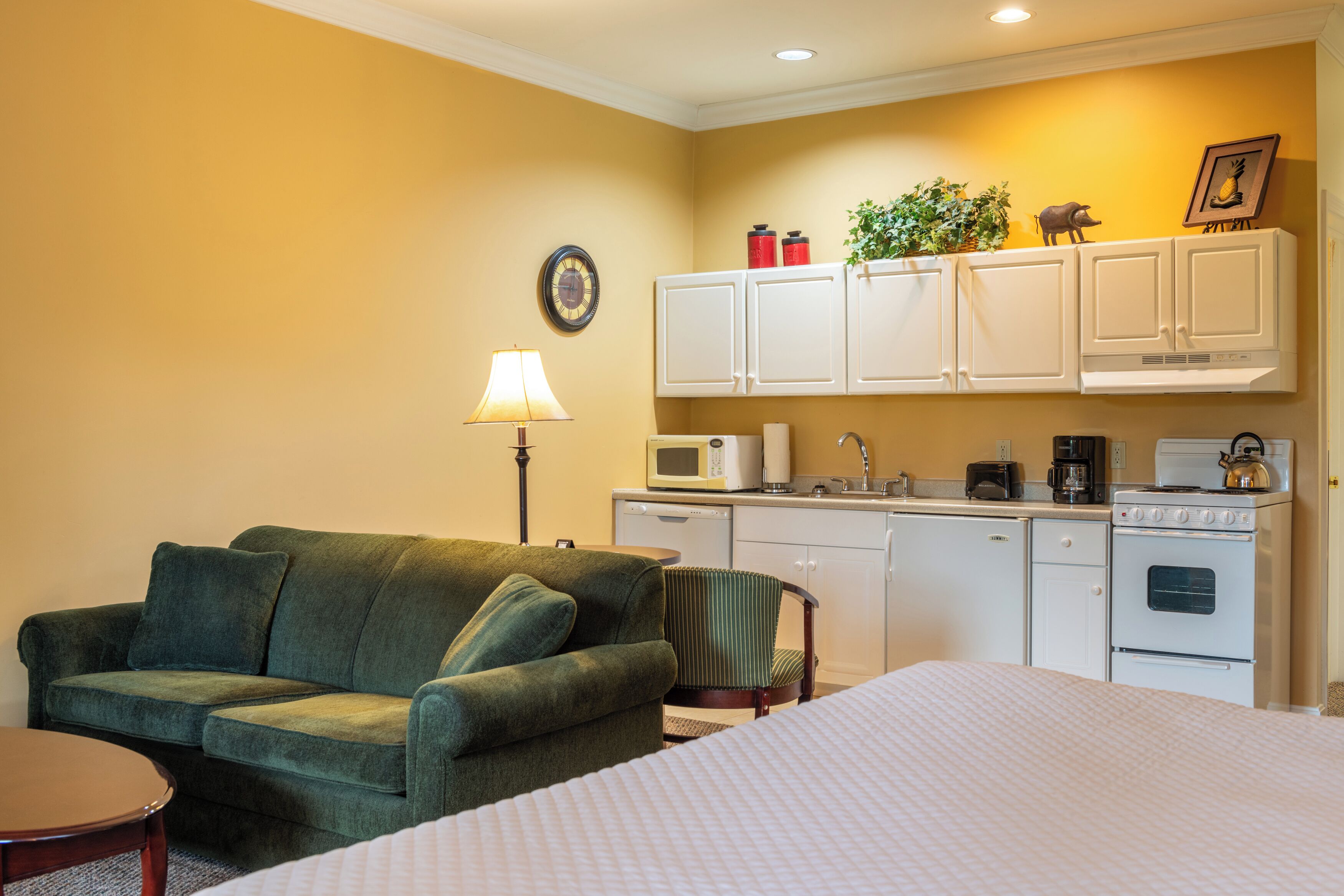 Habitación de hotel compacta con sofá verde, pequeña mesa de comedor, cama con colcha blanca, kitchenette con microondas, tostadora, cafetera y estufa.