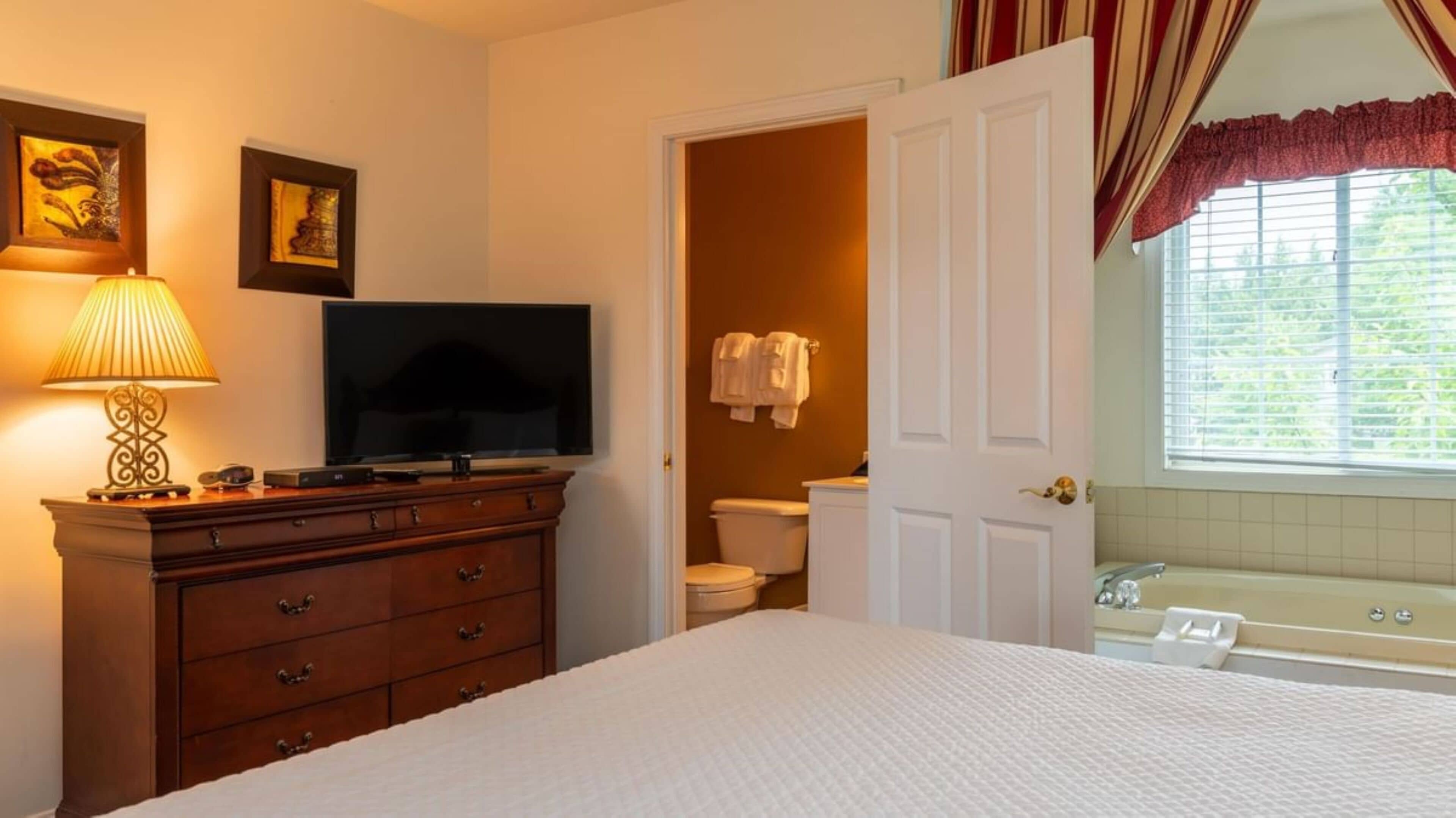 A bedroom with a bed, a dresser, a television, and a bathroom visible through an open door.