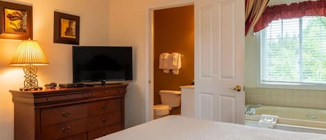 Un dormitorio con cama, cómoda, televisor y baño visible a través de una puerta abierta.