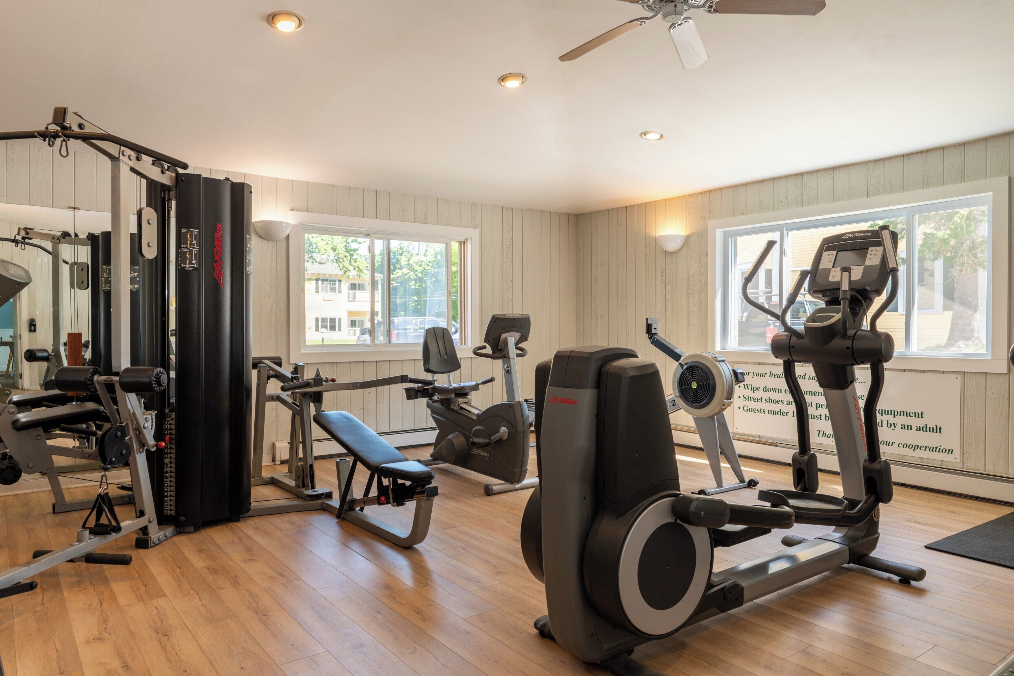 Een sportschool met diverse fitnessapparaten, grote ramen en houten vloeren.