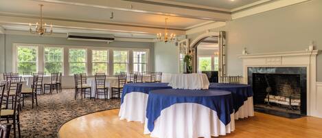 A spacious room with a high ceiling, large windows, and a fireplace. Tables are set up for dining.