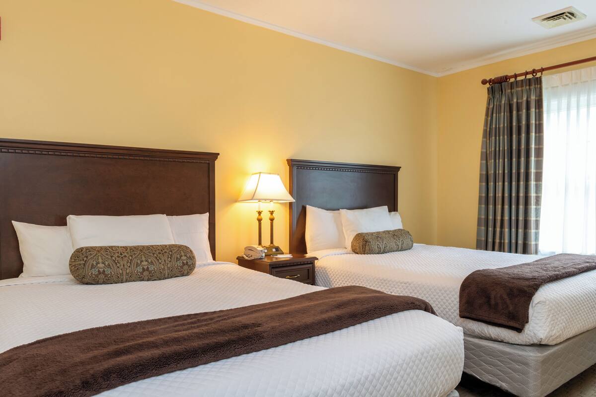 A hotel room with two beds, a nightstand with a lamp, and a window with curtains.