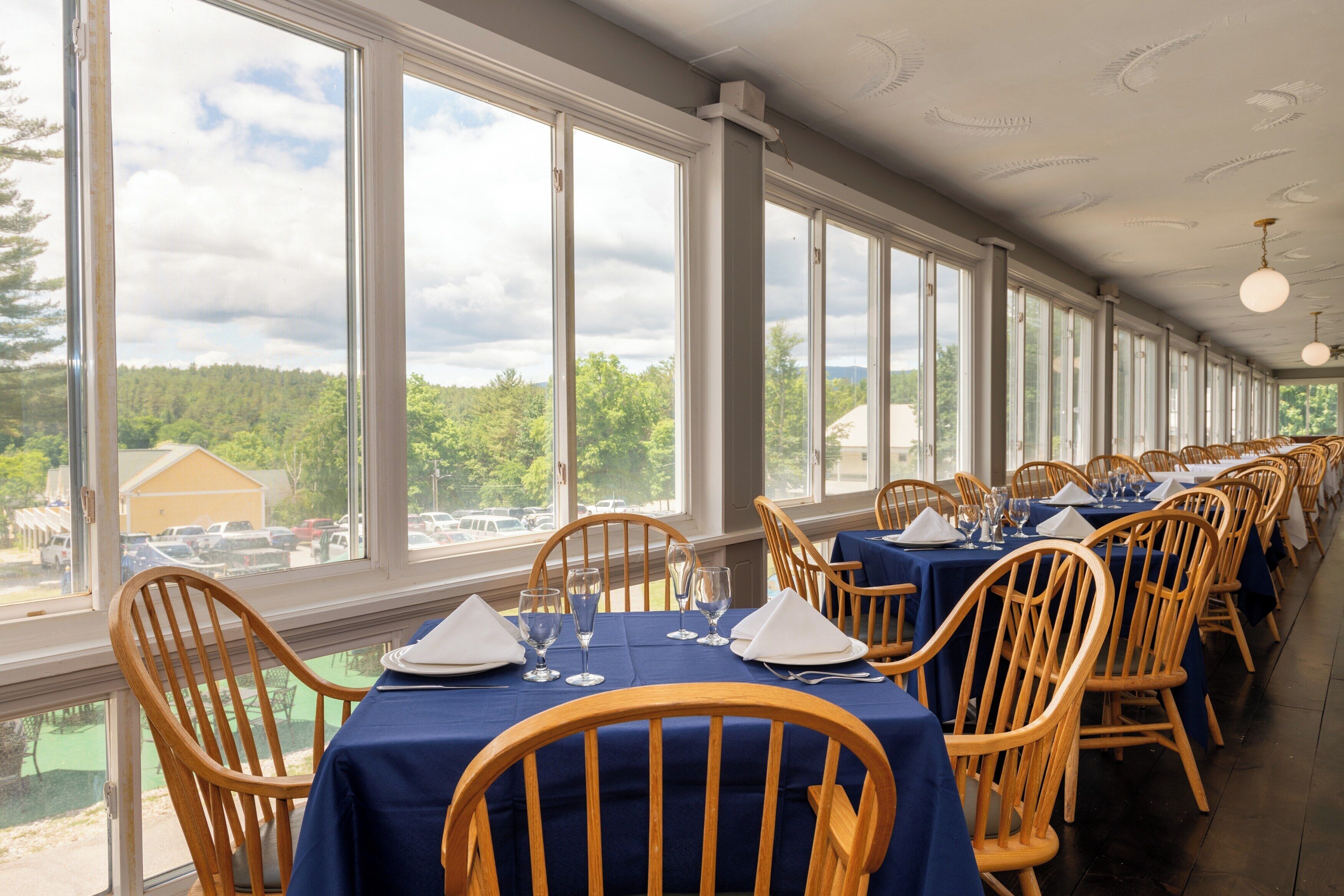 A dining area with tables set for service, large windows with a view of trees and buildings, and wooden chairs.