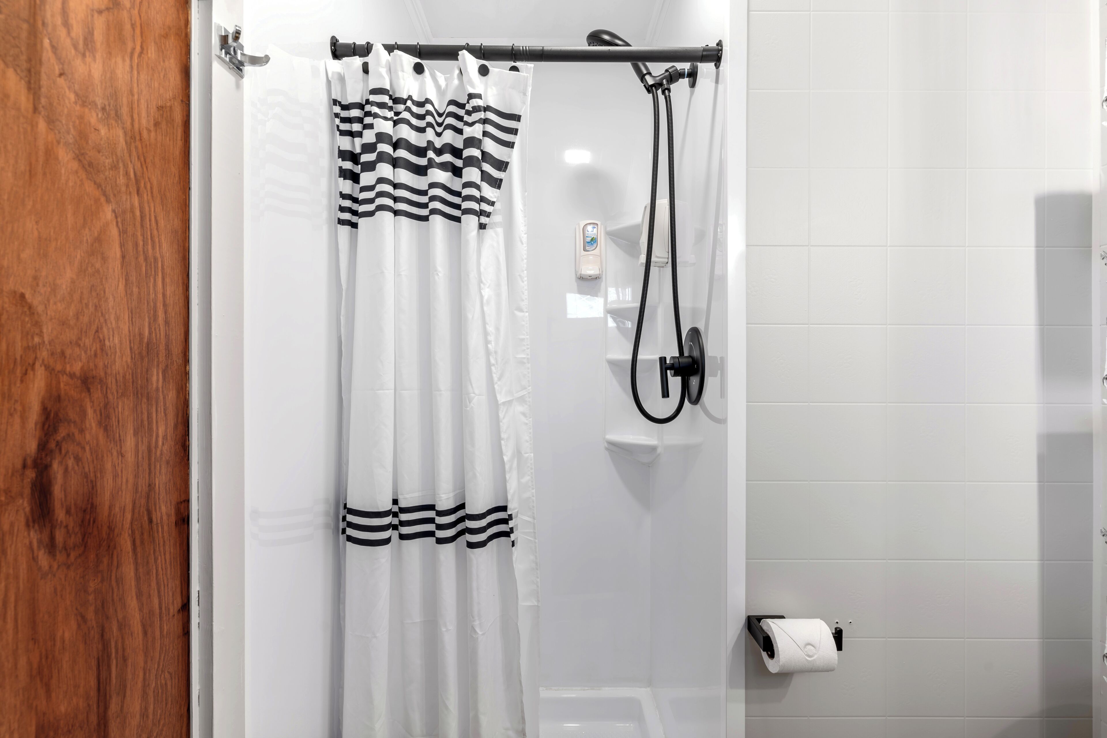 Un baño con cortina de ducha, cabezal de ducha manual y soporte para papel higiénico.