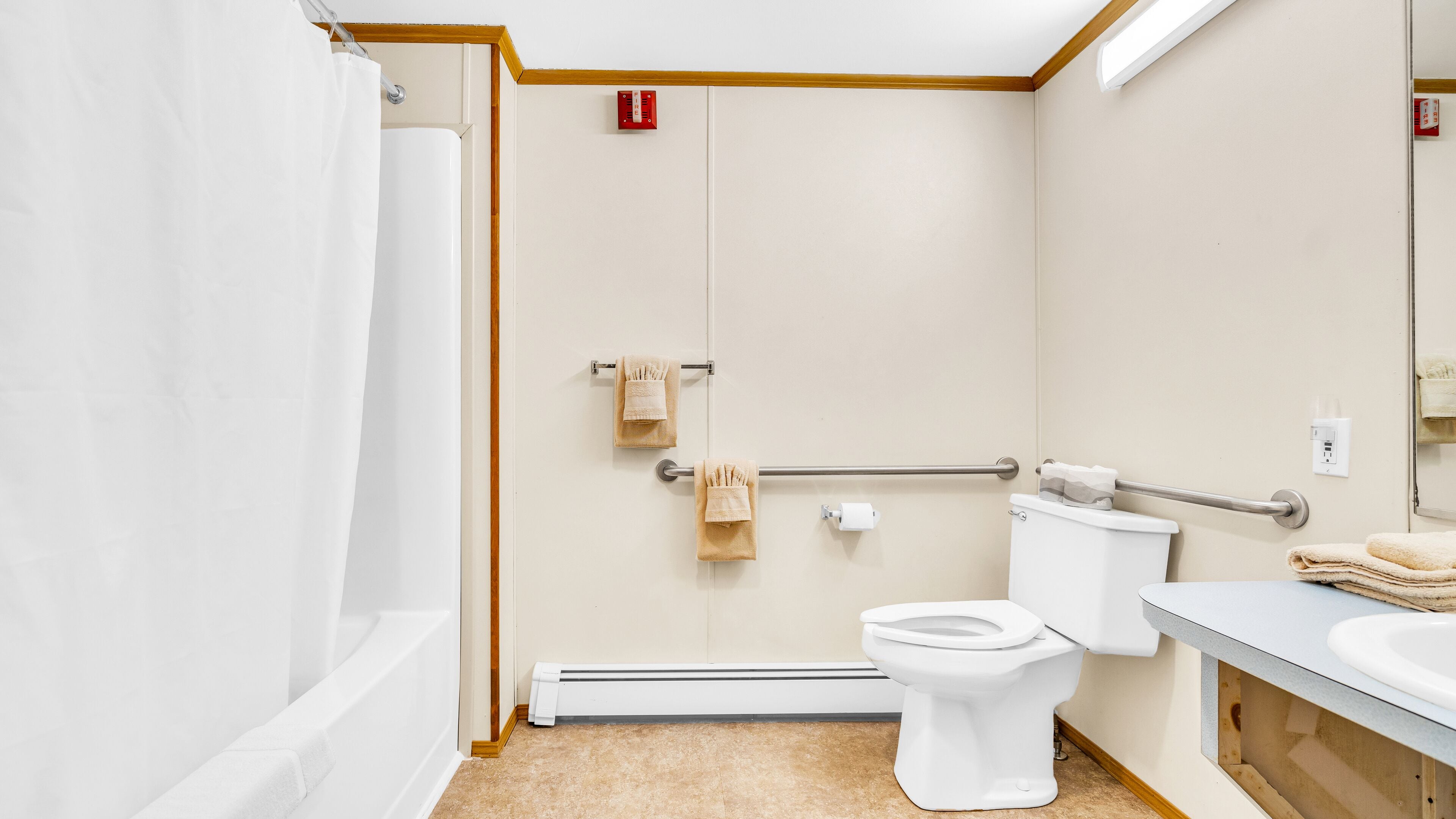 Een badkamer met bad, toilet en wastafel. Er is een handgreep voor toegankelijkheid, een handdoekrek en een rookmelder.