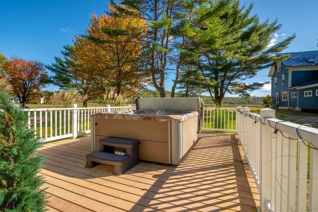 A hot tub on a wooden deck with a white fence, surrounded by trees and a house in the background.