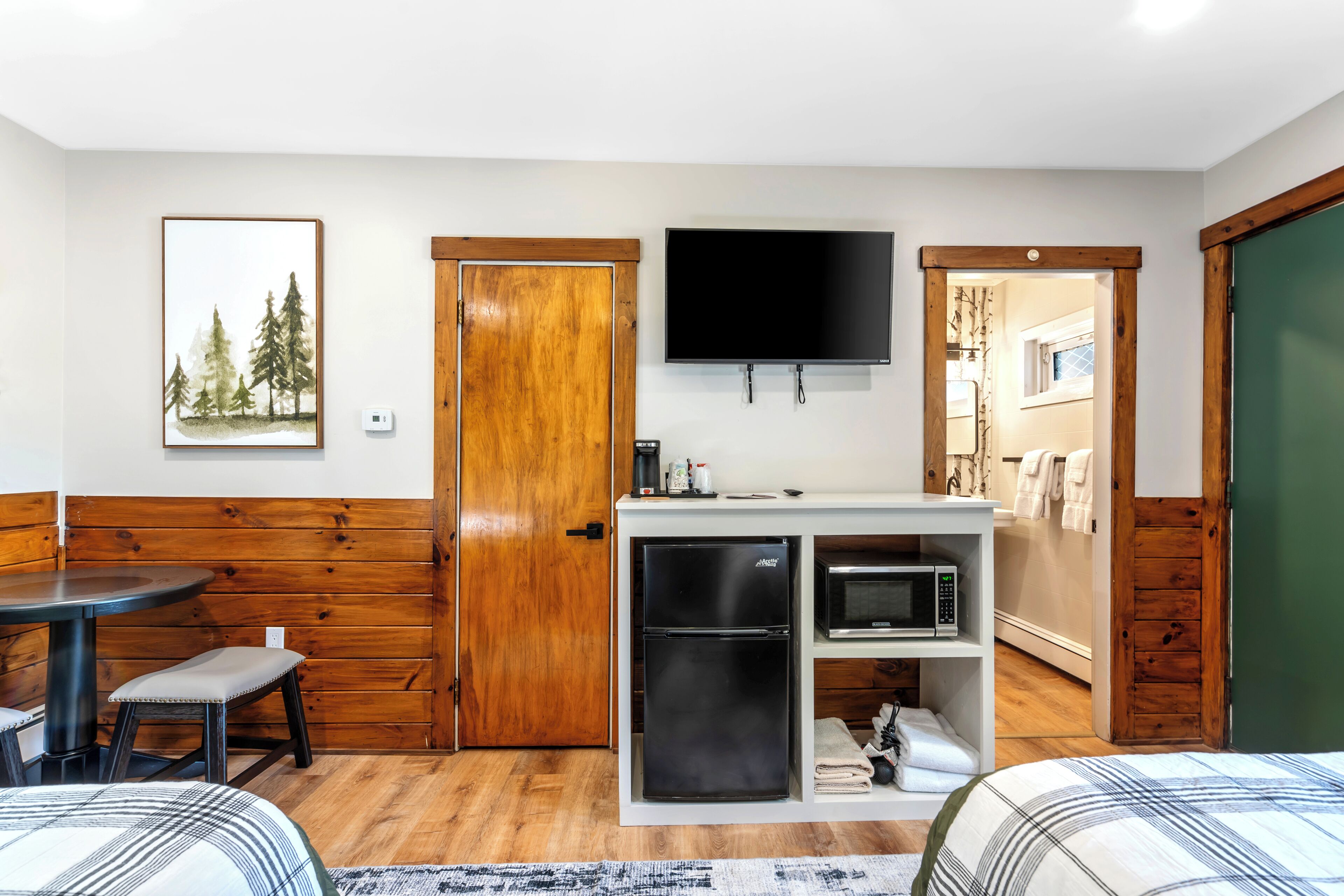Una habitación pequeña con cama, una mesa de comedor, una banca, una kitchenette equipada con refrigerador y microondas, un televisor, y una puerta que da a otra habitación.