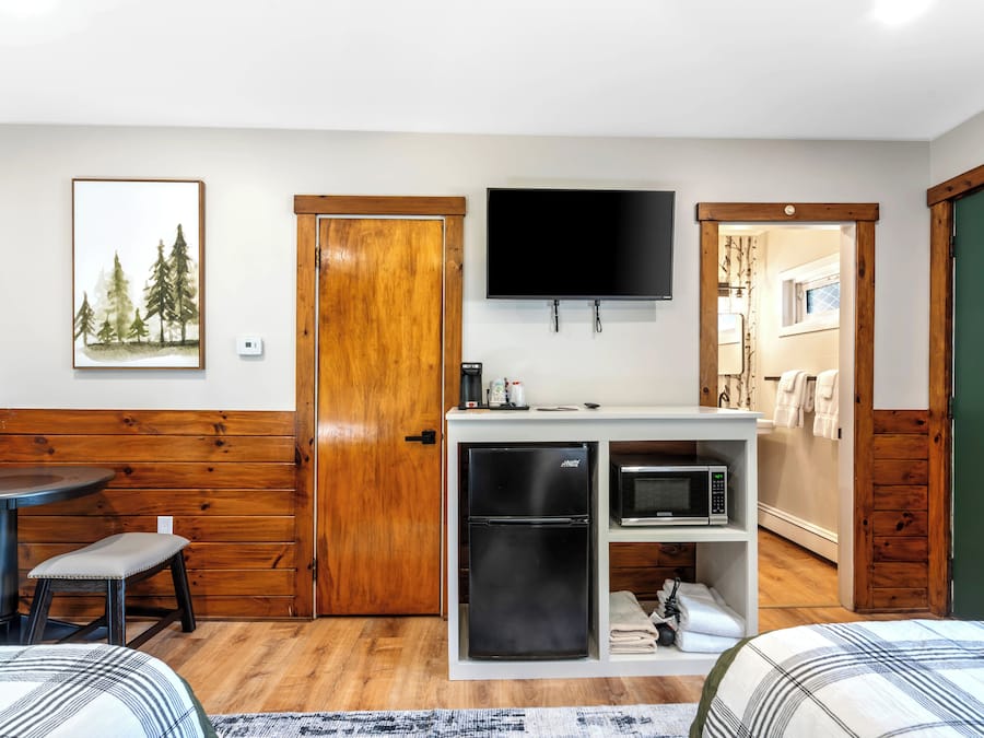 A compact room with a bed, a small dining table, a bench, a kitchenette with a fridge and microwave, a TV, and a door leading to another room.