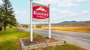 A sign for Rostay Lodging with a mountain logo, located beside a road with a grassy area and trees. - The Inn at the Rostay (Bethel)