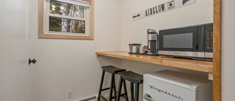 Une cuisine compacte équipée d’un four à micro-ondes, d’une machine à café et d’un réfrigérateur. Une fenêtre offre une vue sur les arbres, et un panneau est fixé au-dessus du comptoir avec l’inscription « AIRGLOW ».