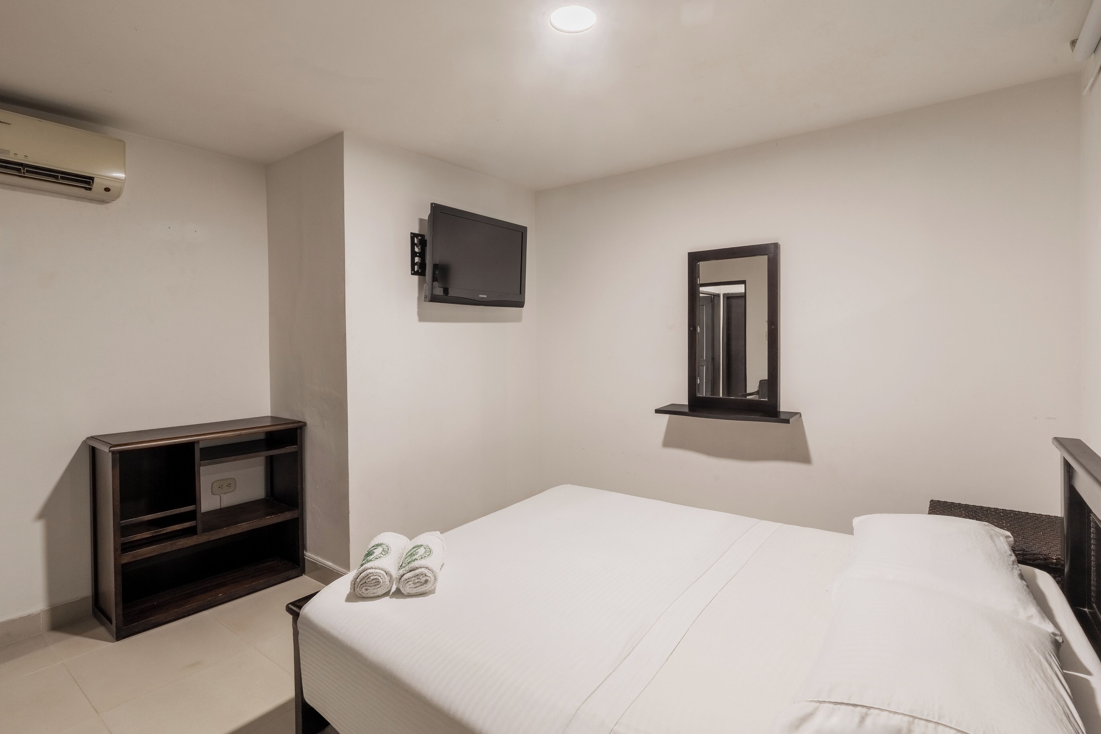 A hotel room with a bed, a TV mounted on the wall, a small shelf, and an air conditioning unit.