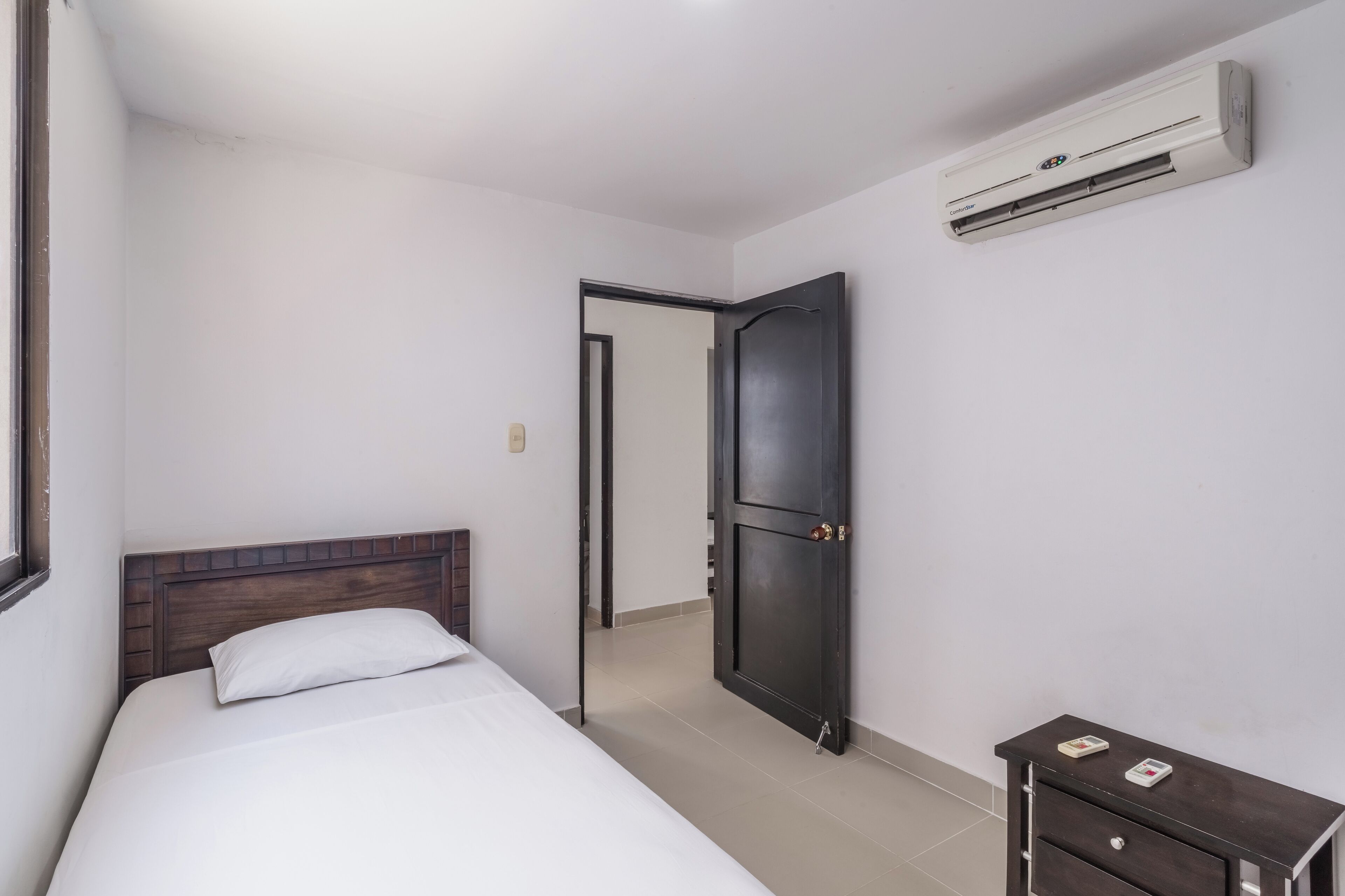 A hotel room with a bed, a dark wooden wardrobe, a small bedside table, and an air conditioning unit mounted on the wall.
