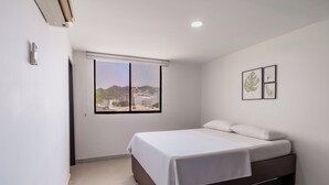 Une chambre à coucher avec un lit, une fenêtre donnant sur des immeubles et des montagnes, et un climatiseur fixé au mur.