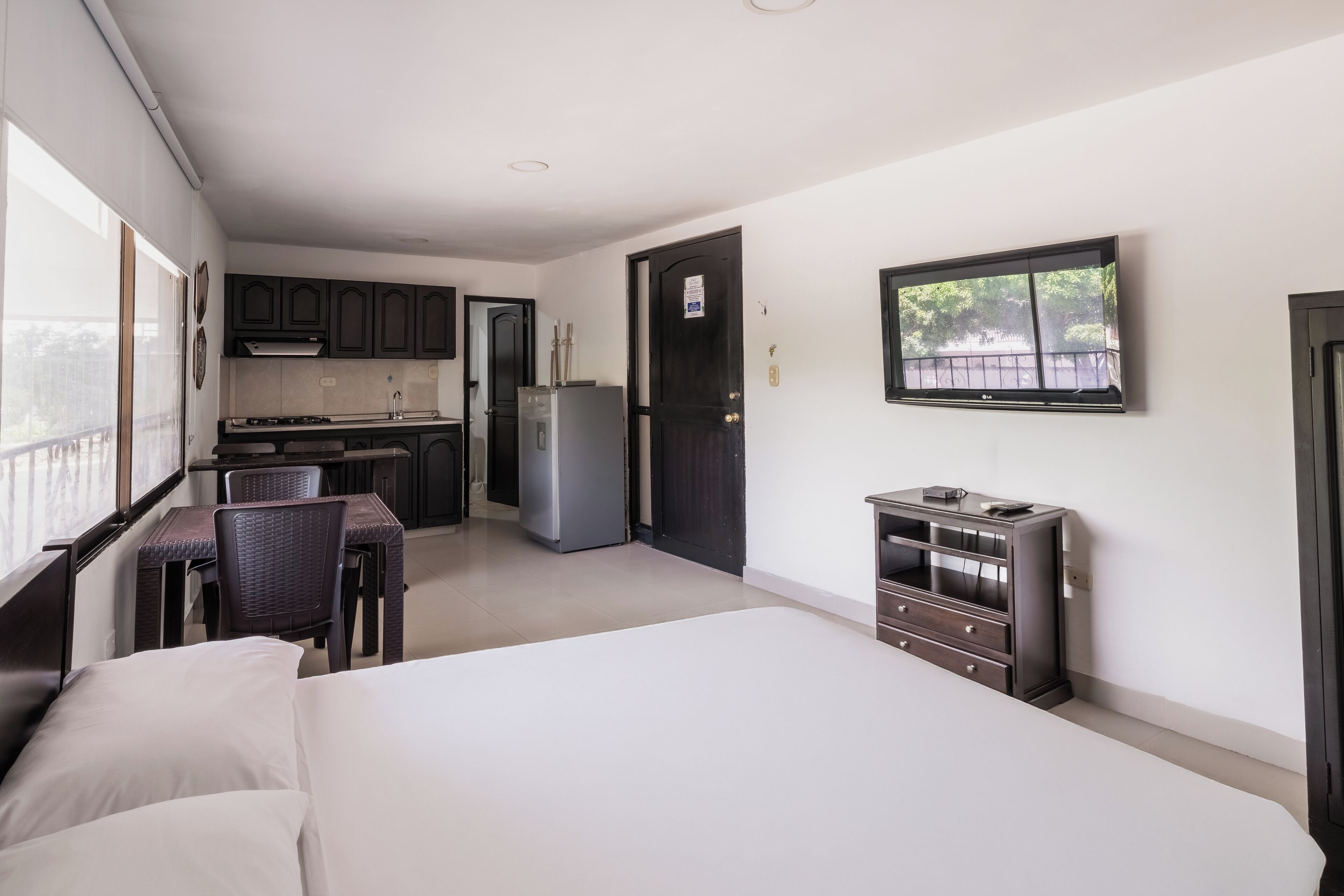 A modern hotel room with a large bed, a desk with a chair, a kitchenette, and a TV mounted on the wall.