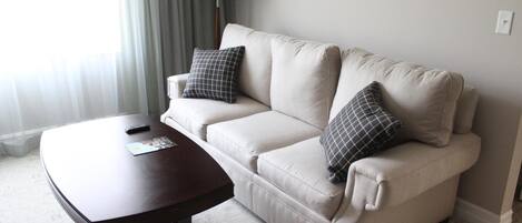 A hotel room with a beige sofa, two matching pillows, a wooden coffee table, and a lamp.