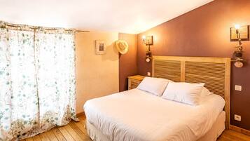Une chambre à coucher avec une tête de lit en bois, un lit recouvert de draps blancs, une table de chevet, une lampe fixée au mur et un rideau orné d’un motif floral.