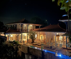 A two-story wooden house with a balcony, outdoor seating area, and a swimming pool.