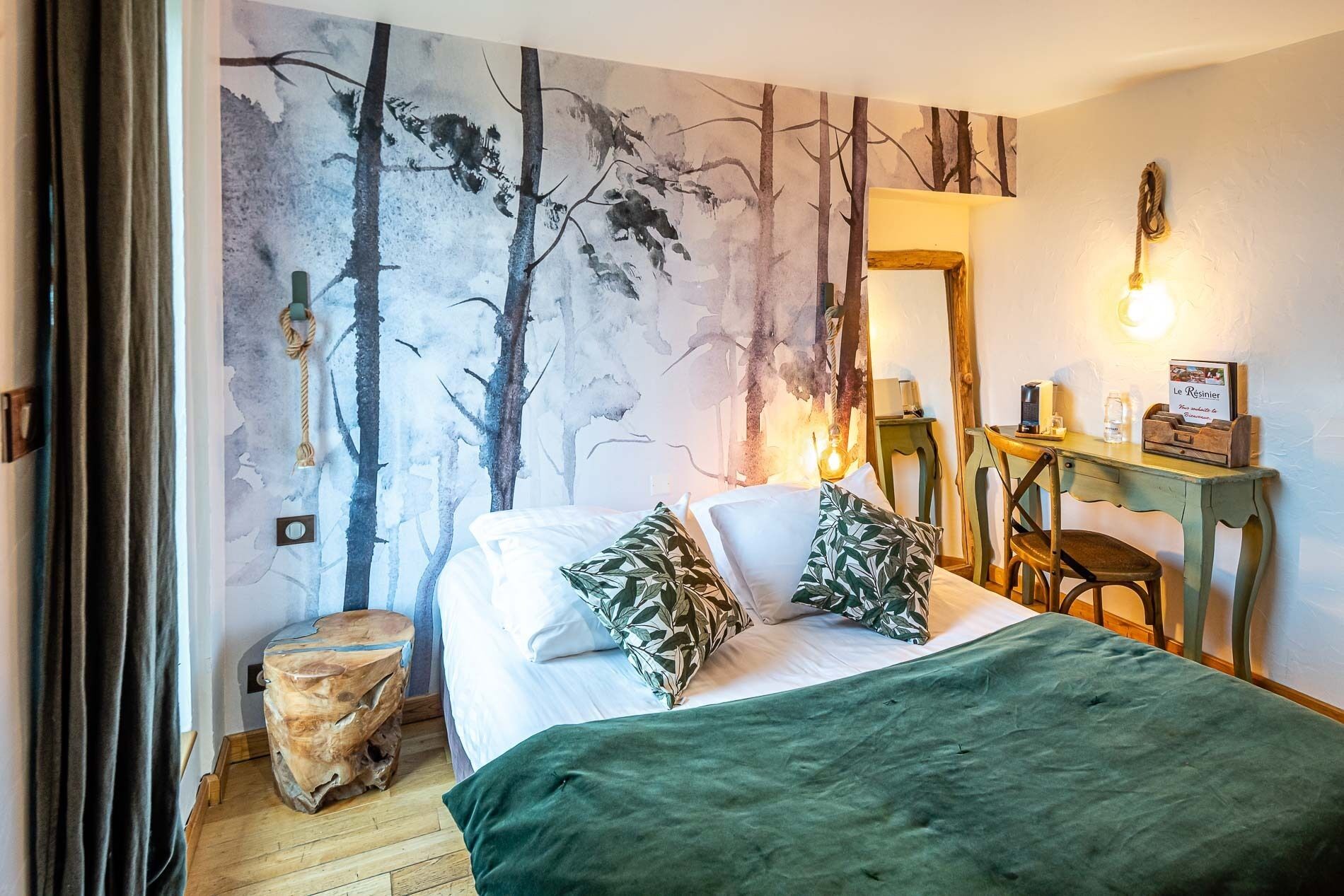 A bedroom with a bed, a desk, a chair, a mirror, and a wall with a tree mural.