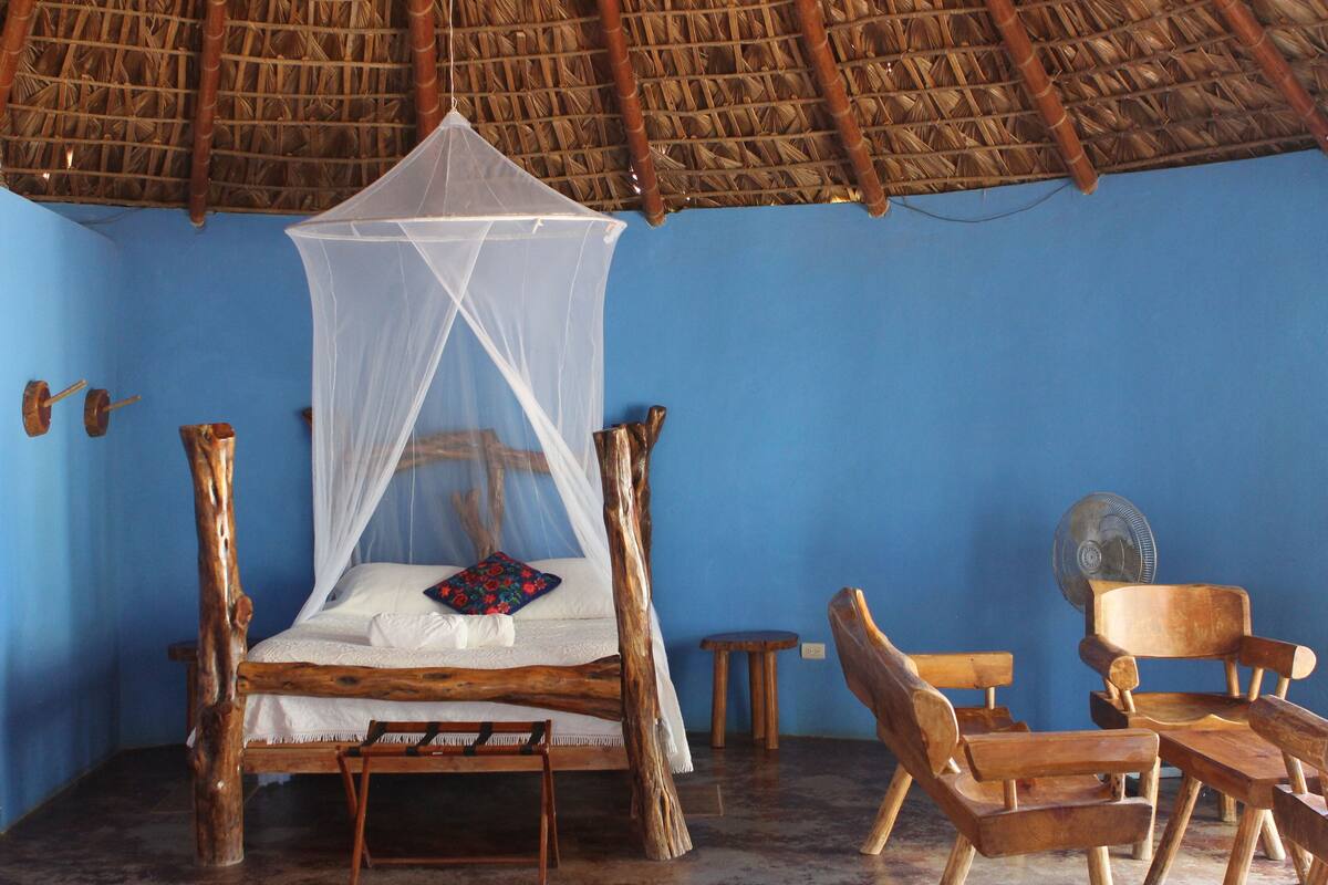 Una habitación con una cama bajo un techo de paja, muebles de madera y una pared azul.