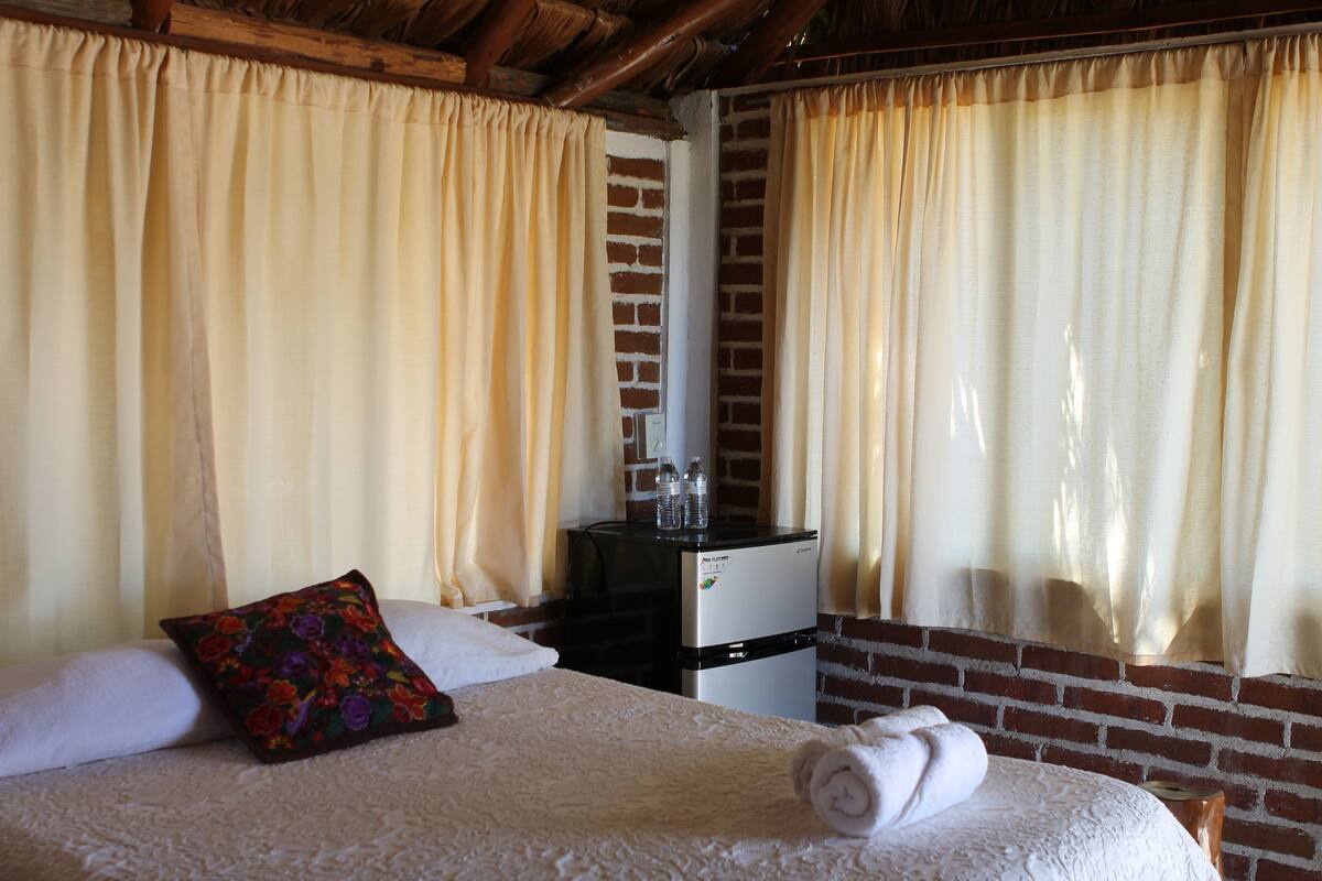 Una habitación de hotel con cama, almohada, toalla enrollada, refrigerador pequeño y cortinas.