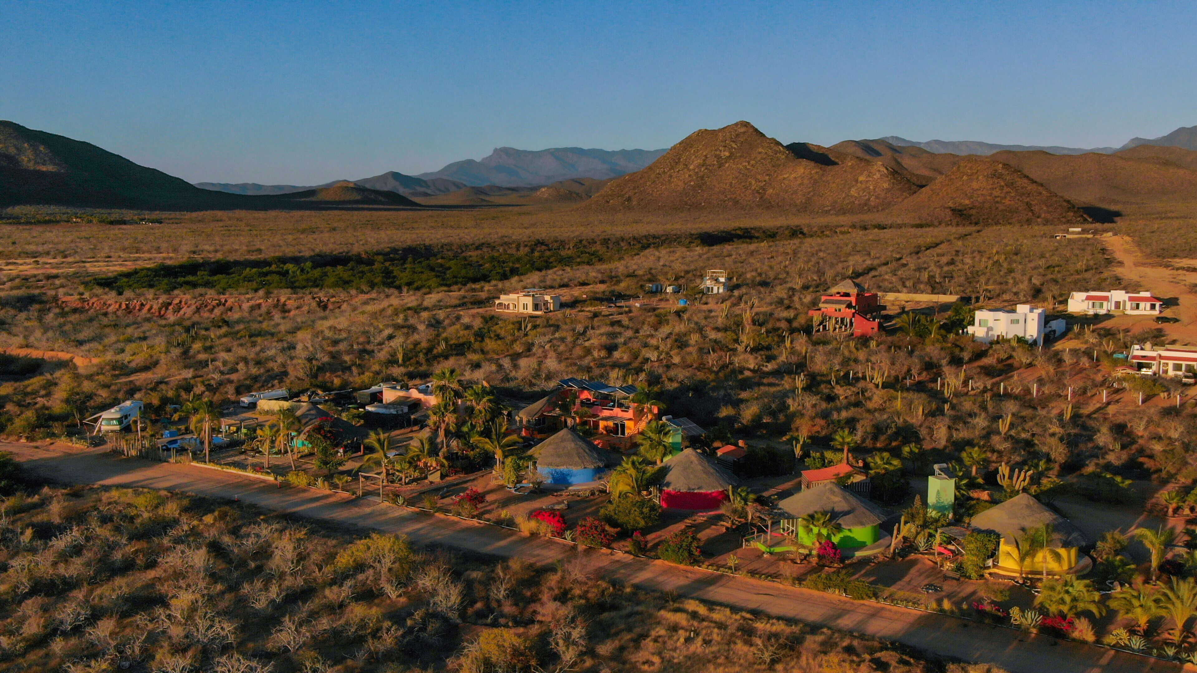 Una comunidad desértica aislada con varias edificaciones, incluyendo casas y una estructura grande, rodeada de montañas y vegetación escasa.