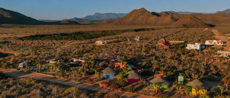 Una comunidad desértica aislada con varias edificaciones, incluyendo casas y una estructura grande, rodeada de montañas y vegetación escasa.