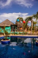 Zona de piscina con un edificio de techo de paja, palmeras y cielo despejado.
