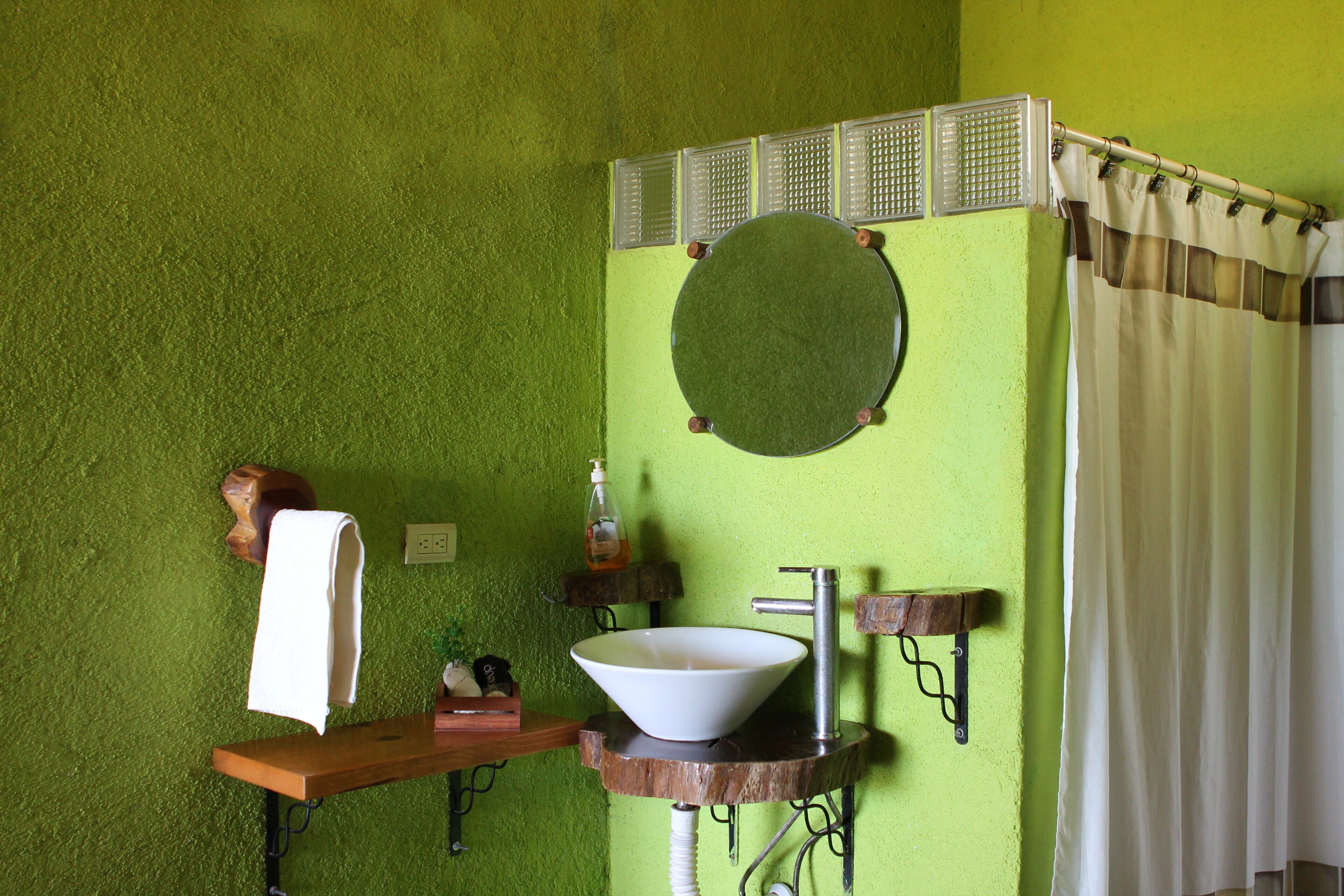 Une salle de bain avec un mur vert, un miroir rond, un lavabo blanc et une étagère en bois.