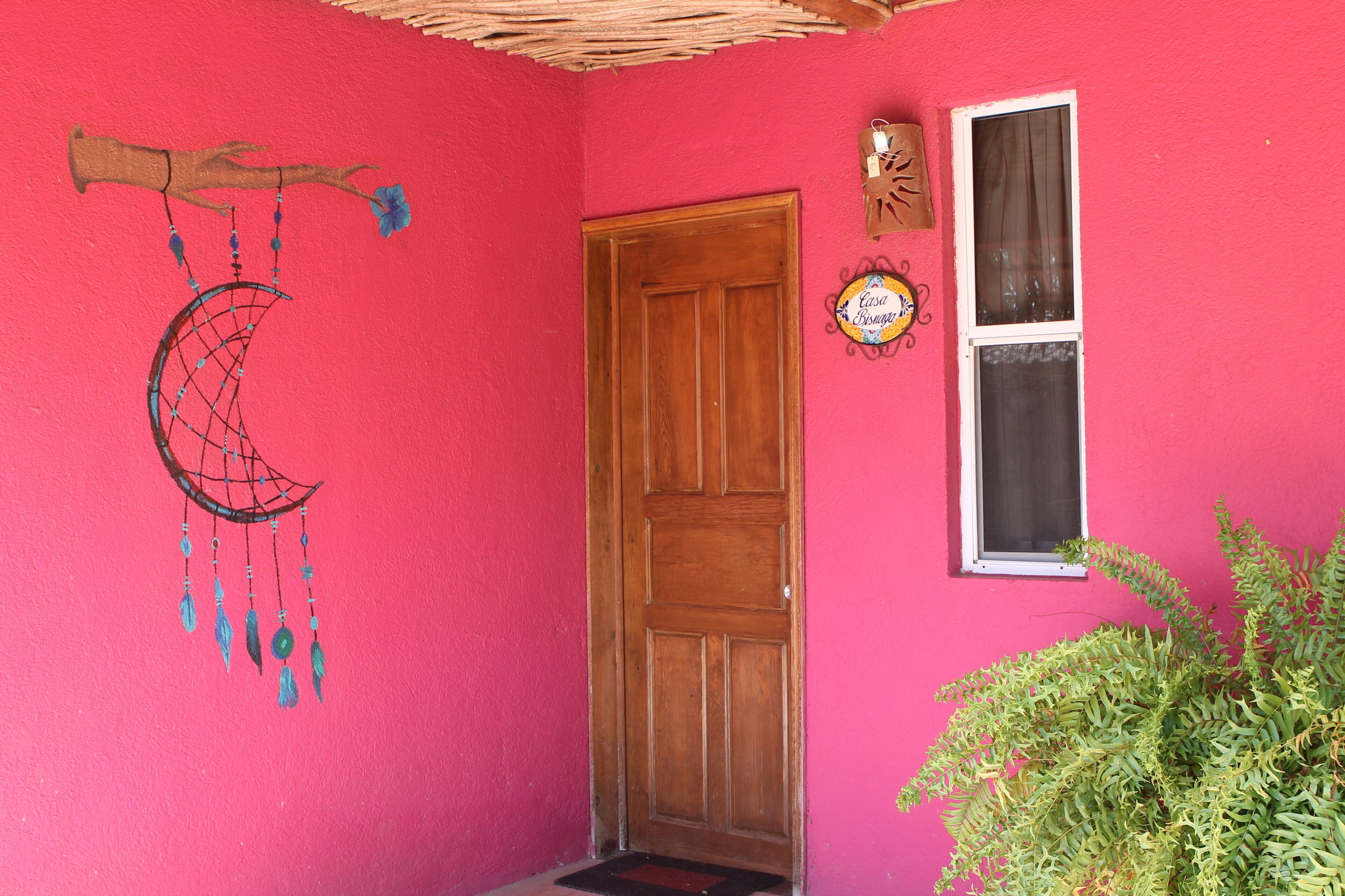 A pink wall with a wooden door, a window, and a dreamcatcher decoration.
