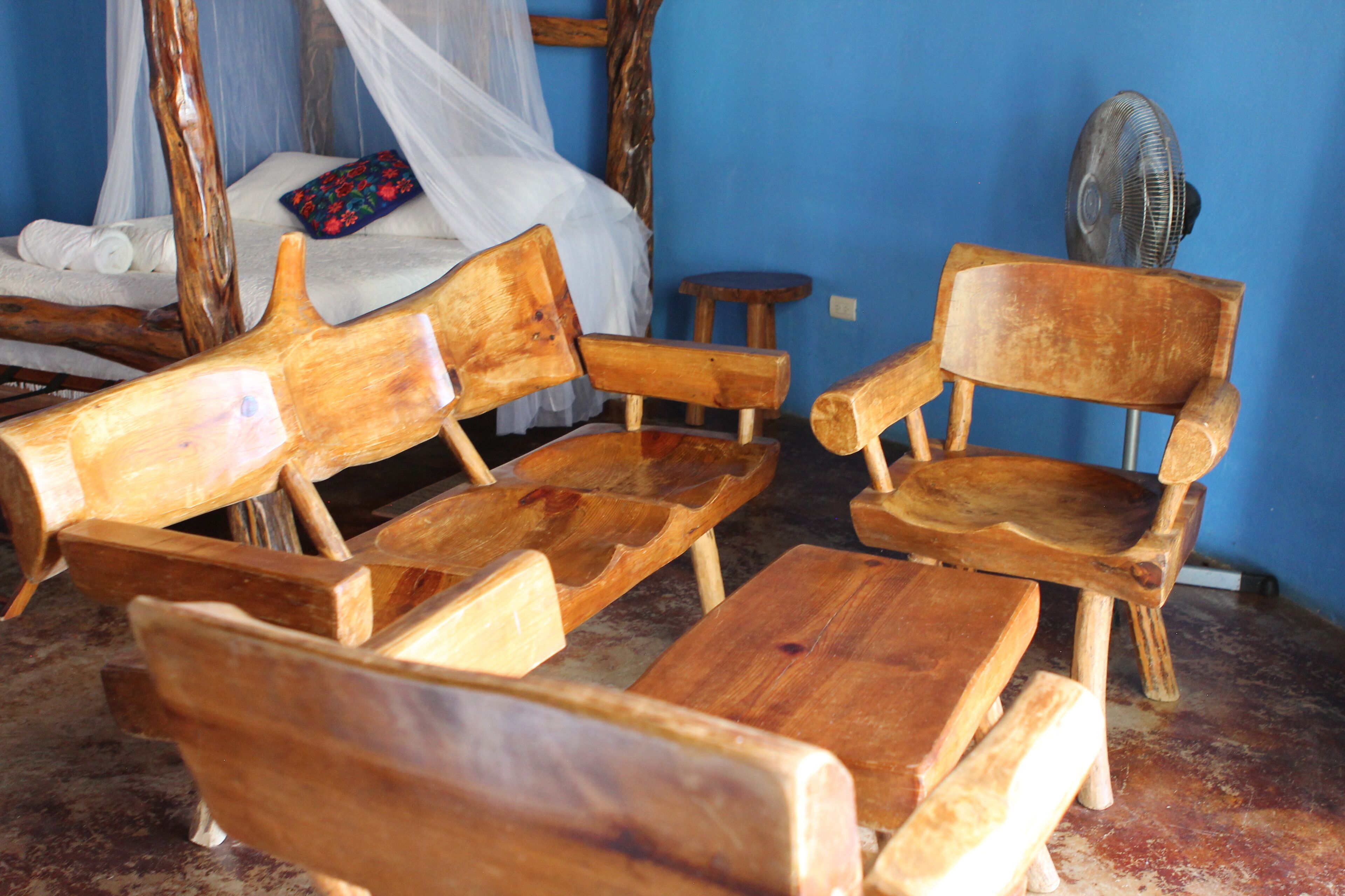 Una habitación con cama con dosel, una silla de madera, un taburete pequeño y un ventilador.