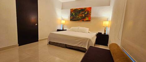 A hotel room with a bed, bedside tables, a bench, a ceiling fan, and a painting on the wall.