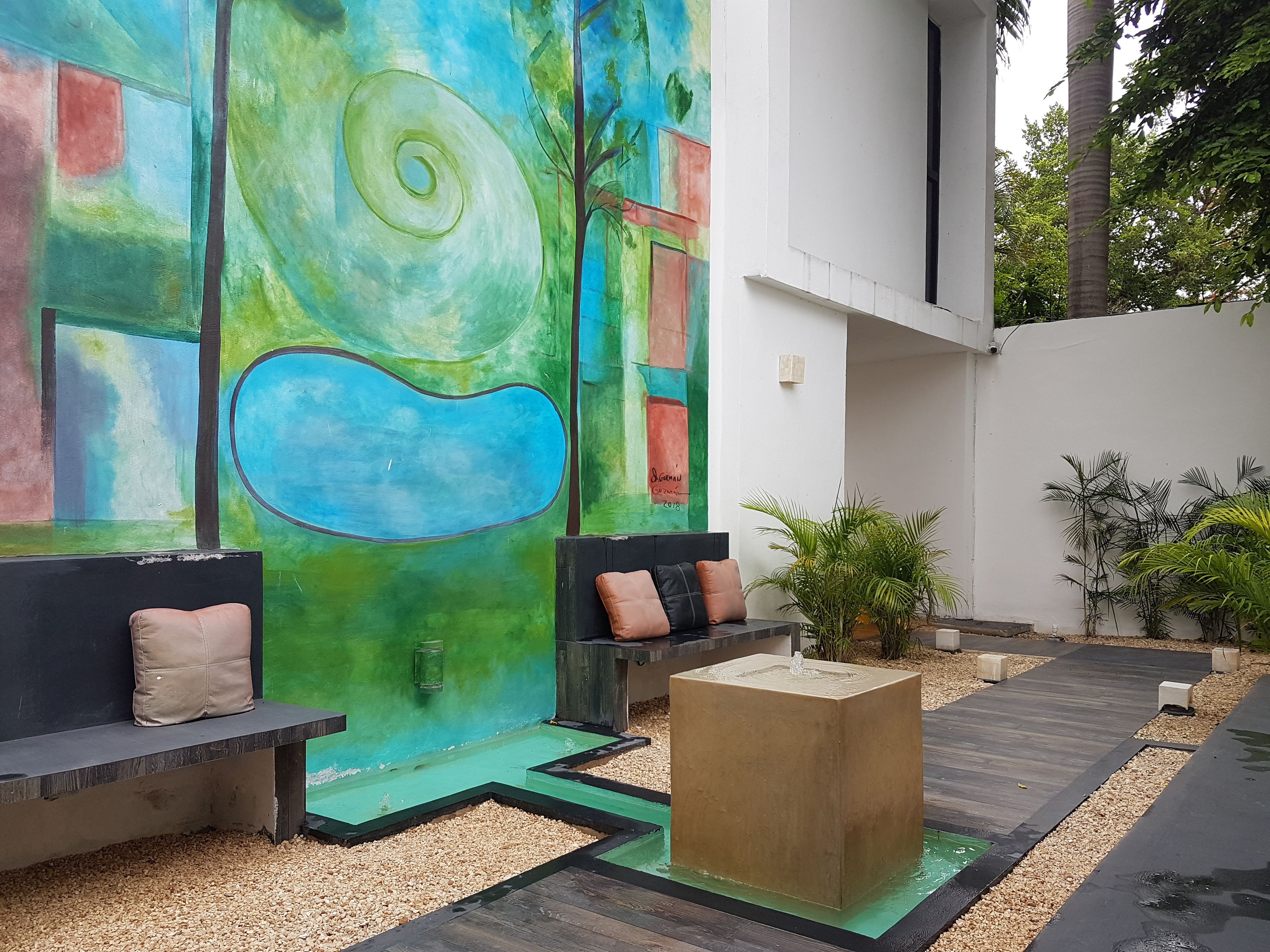 Un área de estar exterior moderna con un mural abstracto de colores, una fuente de agua y una terraza de madera.