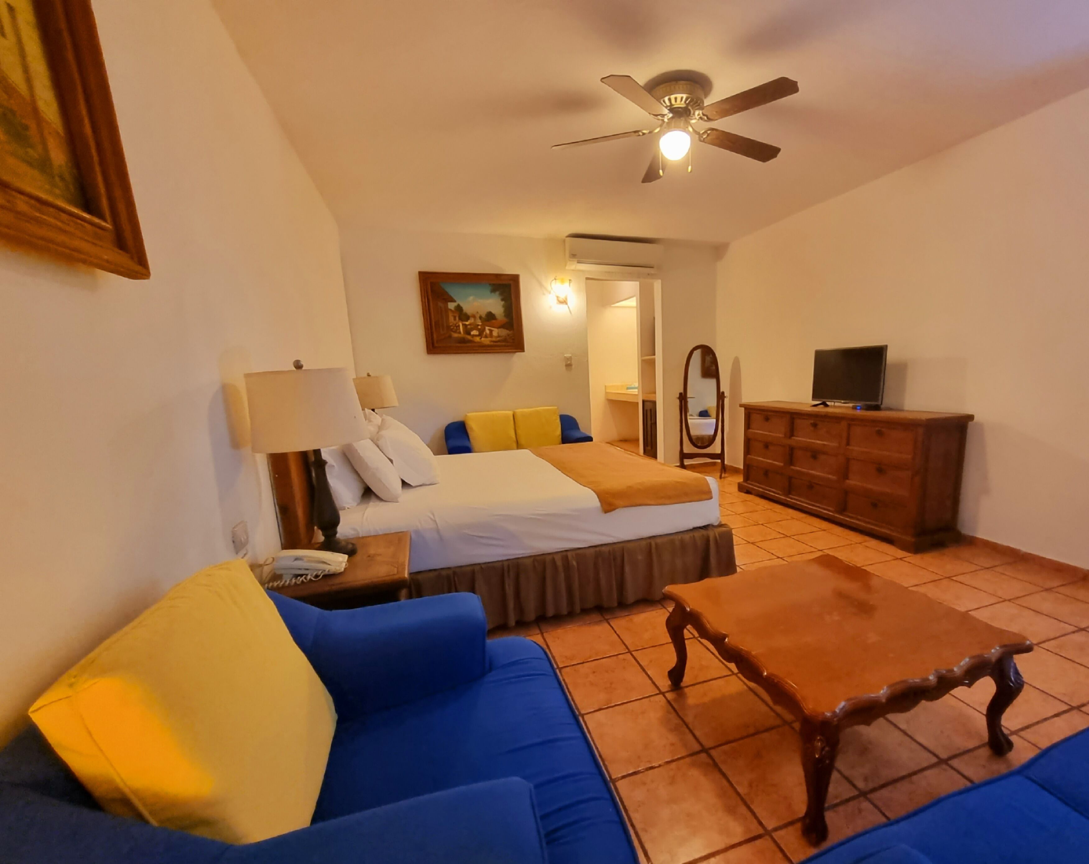 Une chambre d’hôtel comprenant un lit, un canapé, une table basse en bois, une télévision, une commode, un ventilateur de plafond et un tableau accroché au mur.