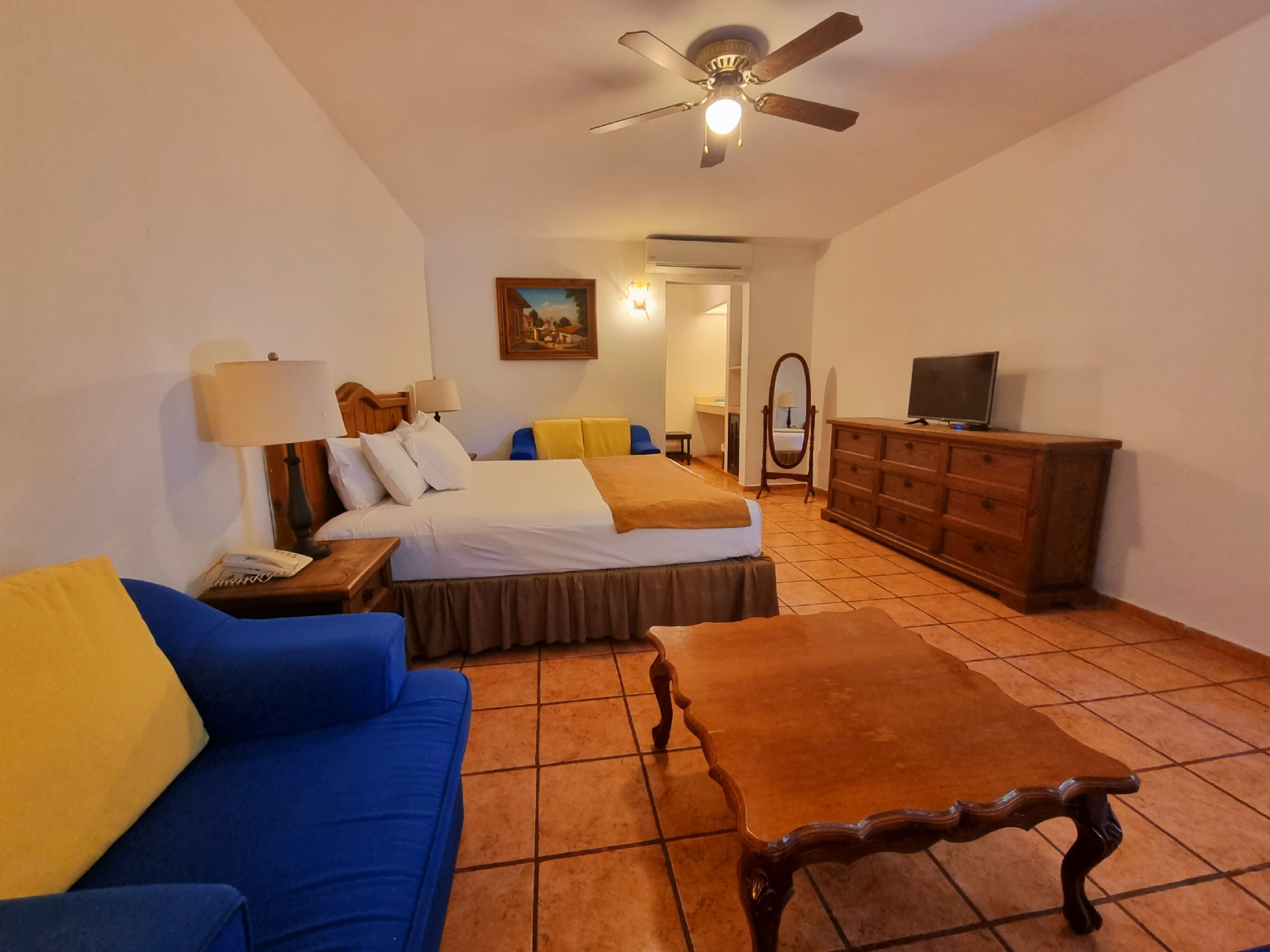 A hotel room with a bed, a blue sofa, a wooden table, a television on a dresser, and a ceiling fan.