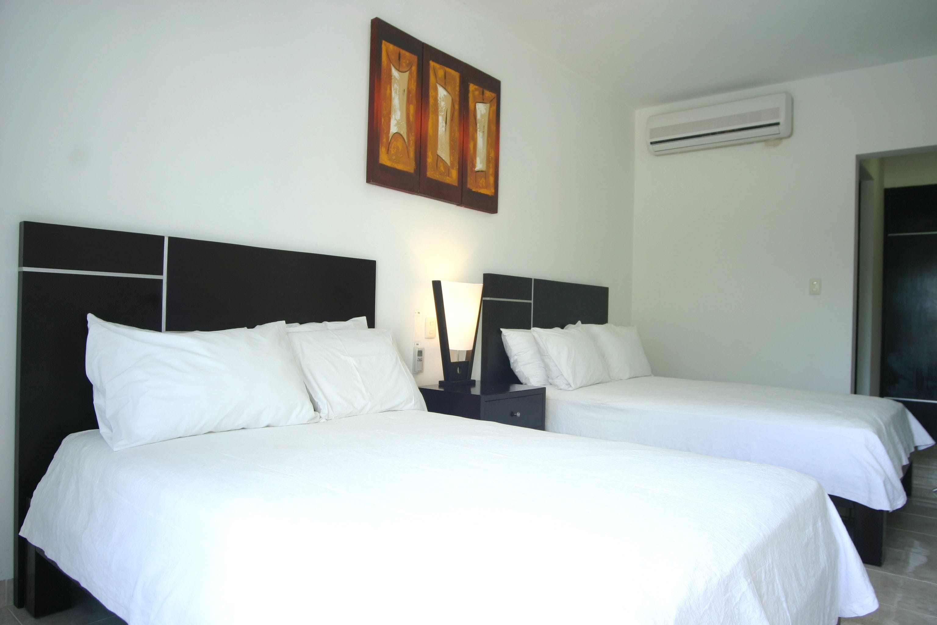 A hotel room with two single beds, a nightstand with a lamp, a wall-mounted air conditioner, and a door leading to another room.
